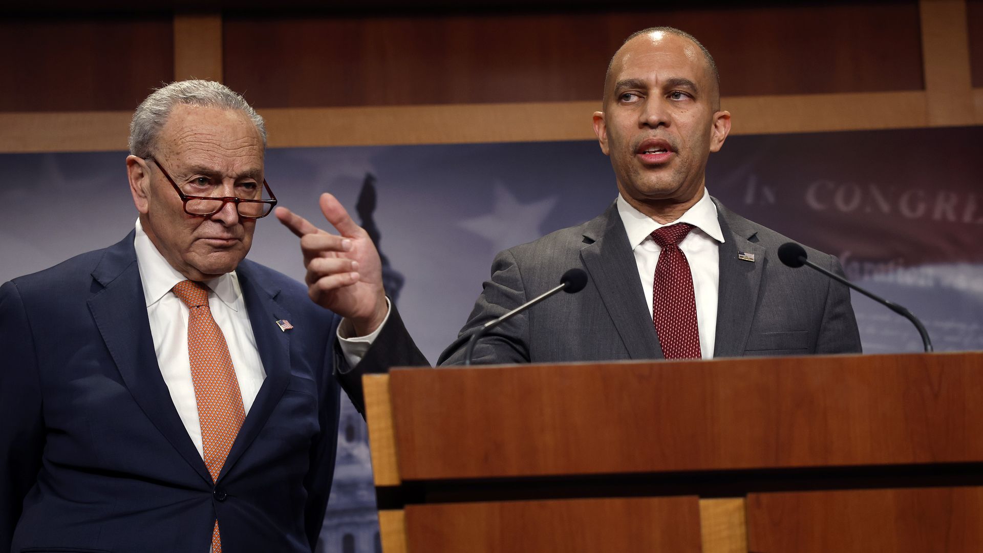 Sen. Chuck Schumer, wearing a blue suit, and Hakeem Jeffries, wearing a gray suit and speaking at a wooden podium.
