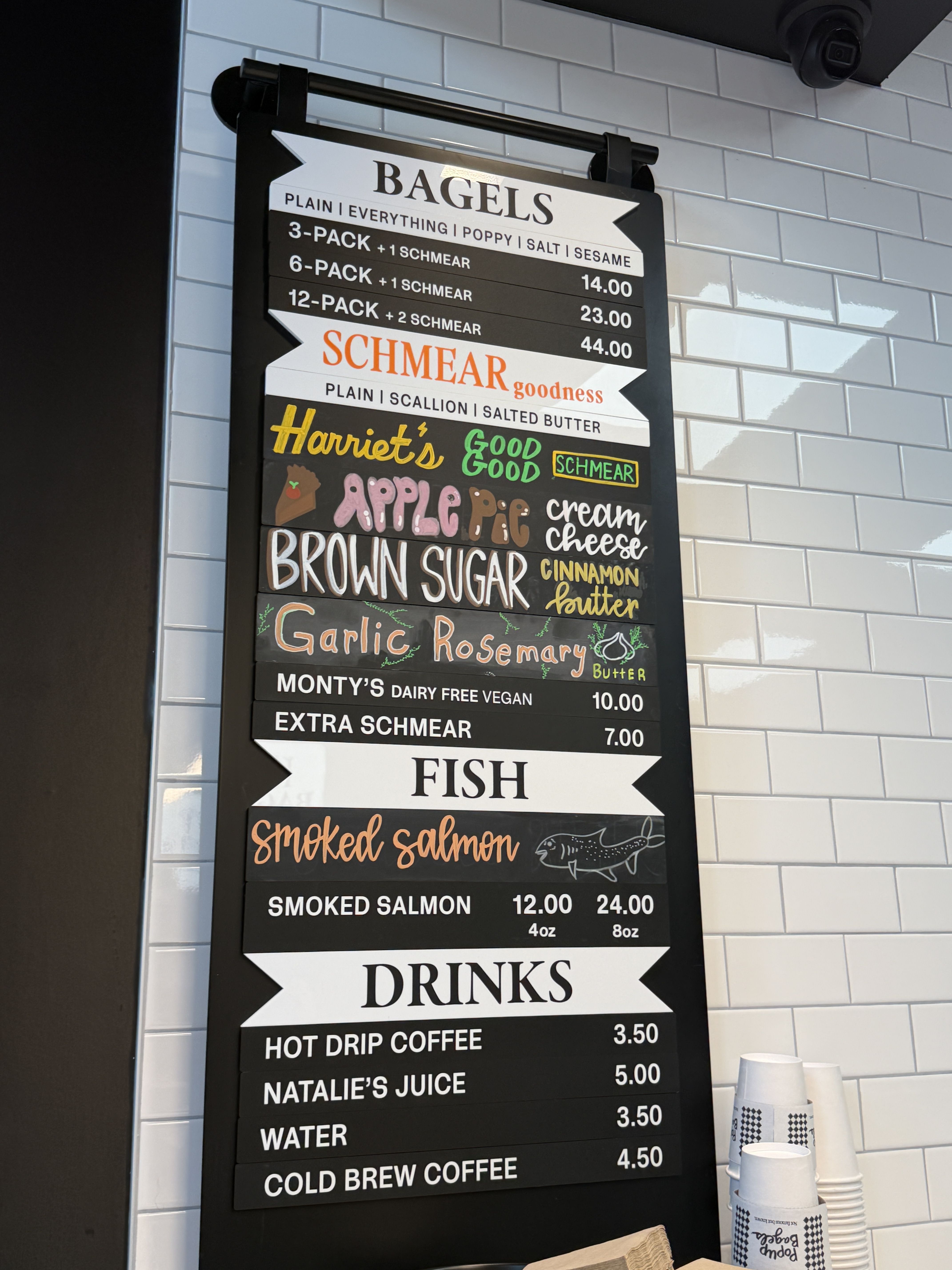 Tall chalkboard menu on a white tiled wall advertising BAGELS with prices and schmear options; includes colorful flavor sections and a stack of cups at the bottom right.