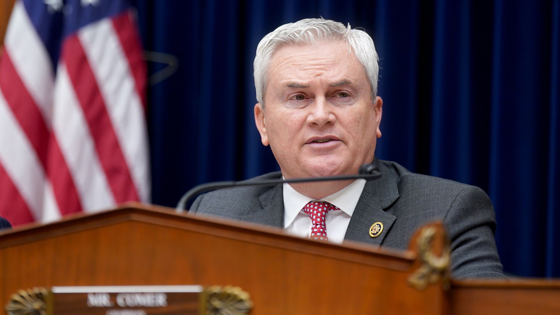 Oversight Committee Chair James Comer, wearing a gray suit, white shirt and red tie, sitting behind a committee dais in front of blue drapes and an American flag.