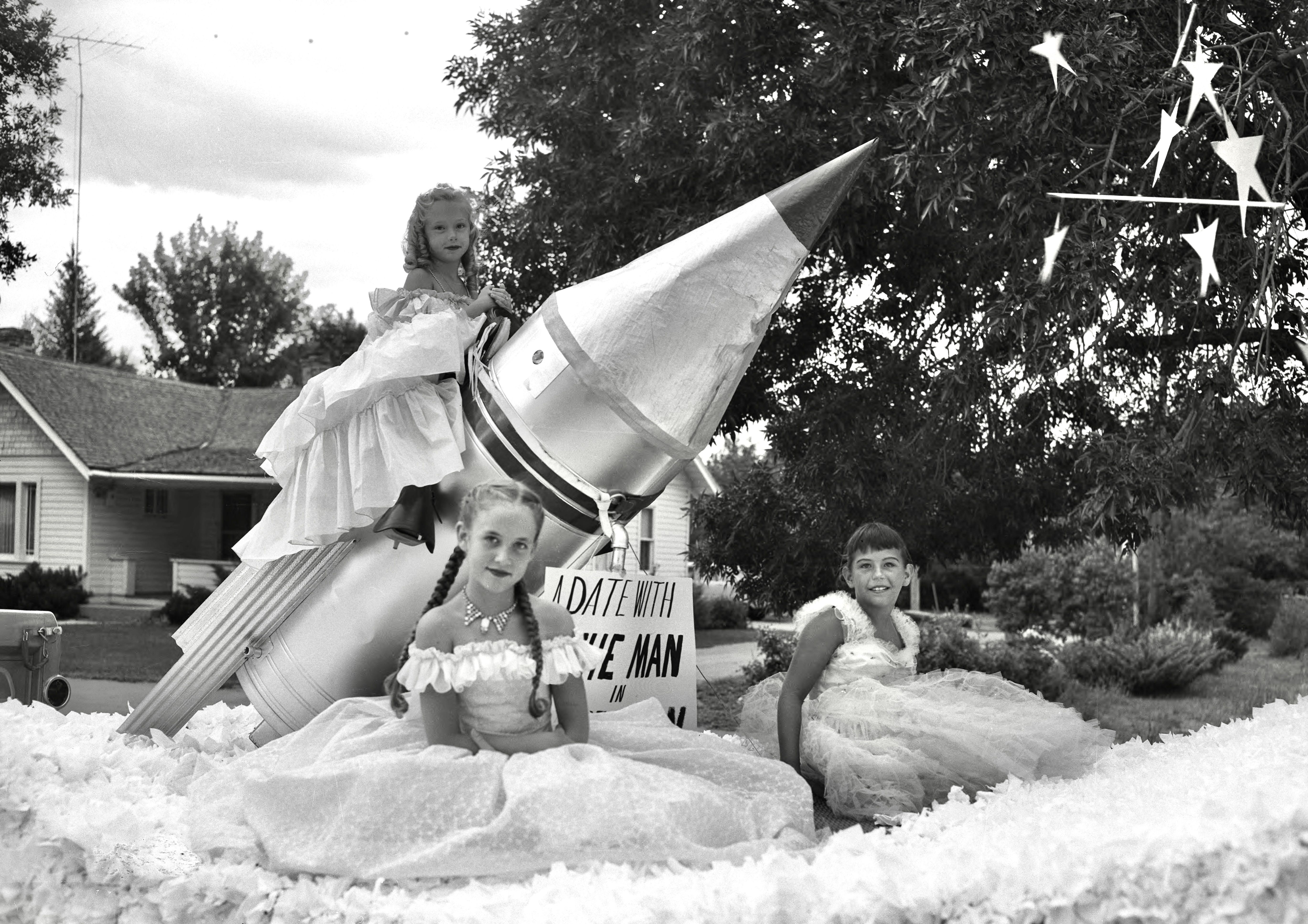 Three girls pose on a parade float featuring a rocket and a sign that reads "A date with the man on the moon."