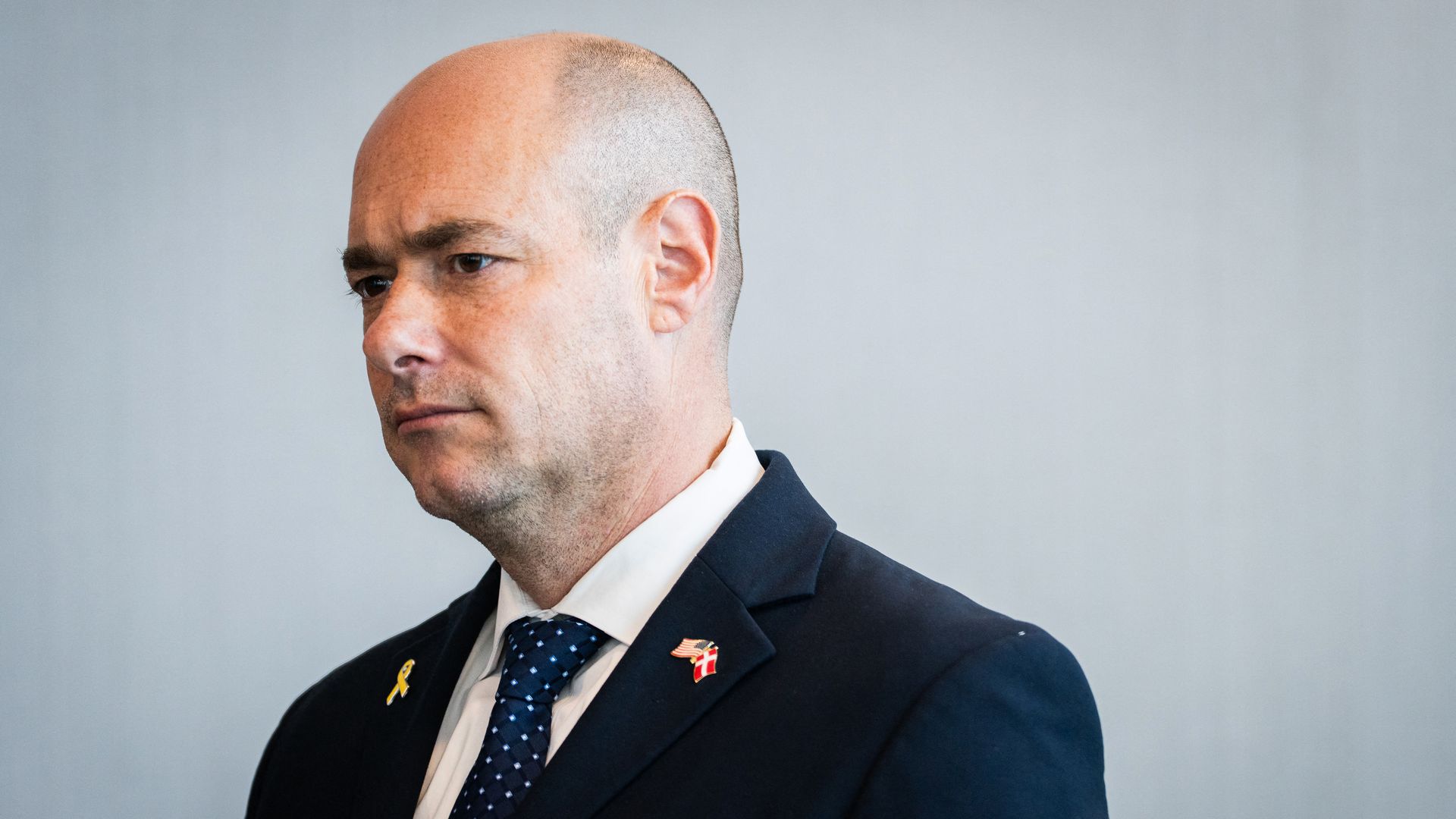 Rep. Greg Landsman, wearing a blue suit with pins on his lapels and standing in front of a light gray wall.