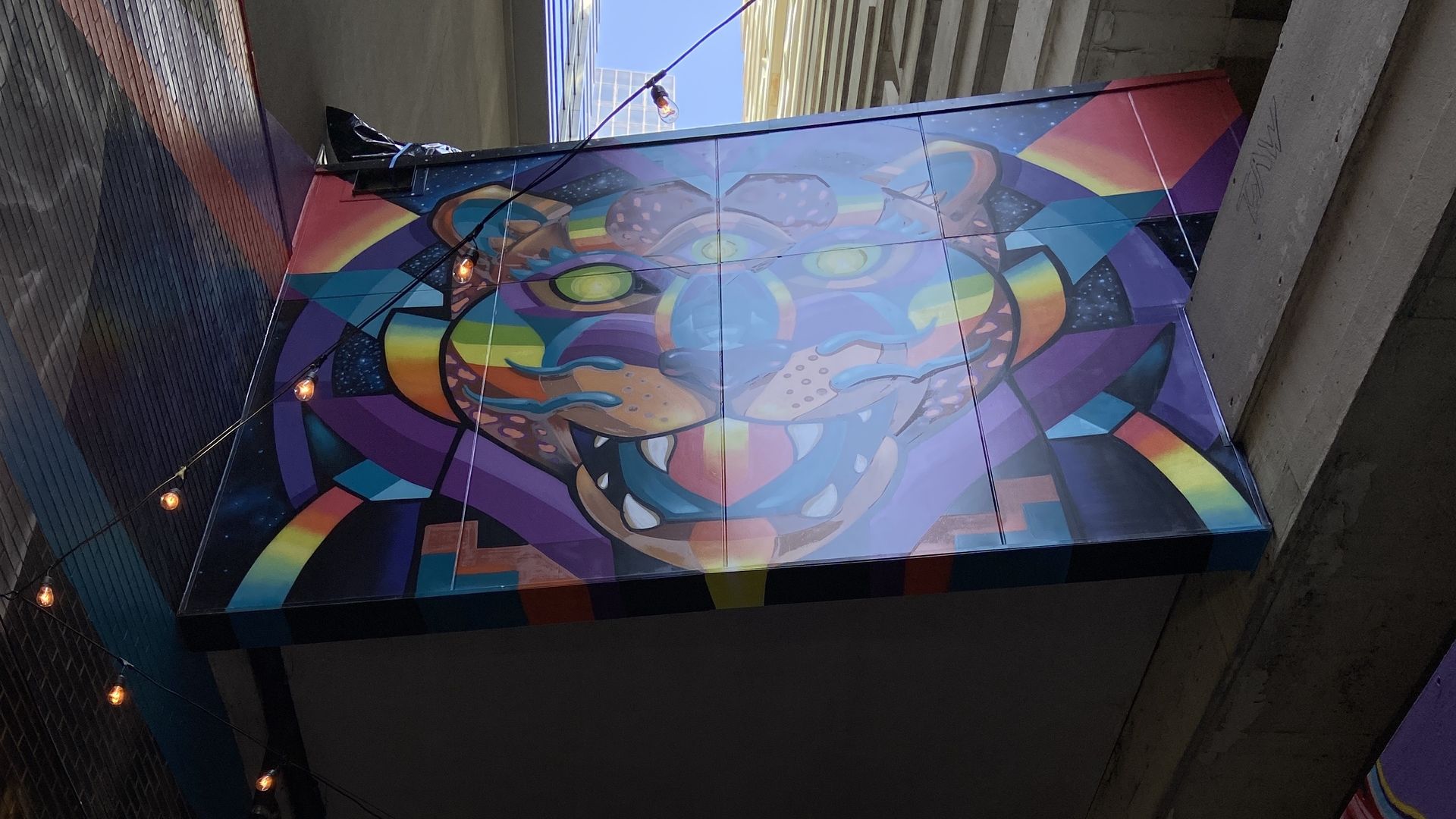 Vibrant geometric tiger mural on a suspended rectangular panel between tall buildings in an urban alley. Bright blue, purple, yellow, and green shapes; string lights and a clear blue sky above.