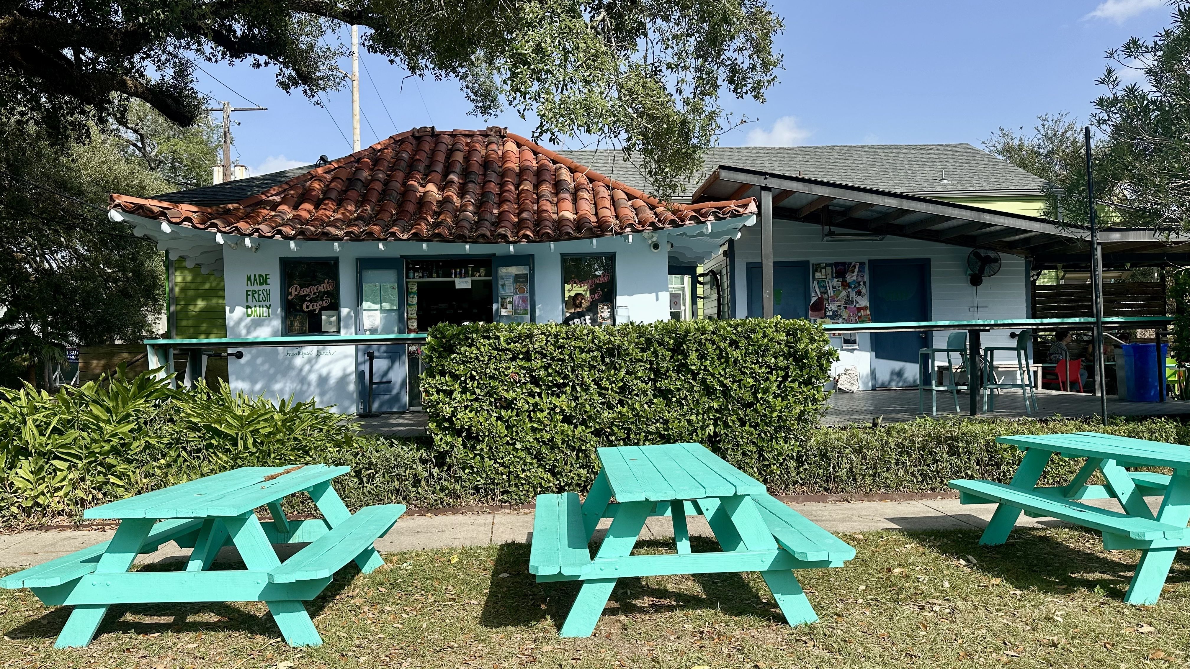 Blue building with terracotta roof housing Pagoda Cafe, surrounded by greenery, and three bright turquoise picnic tables on grass under a clear blue sky.