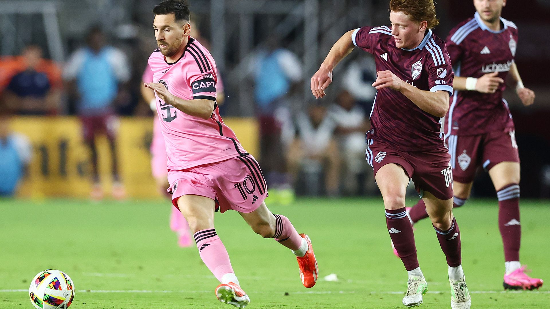 Soccer match: a player in a pink kit runs toward the ball while a maroon-clad opponent chases from the right; another maroon player is in the background on a green pitch.