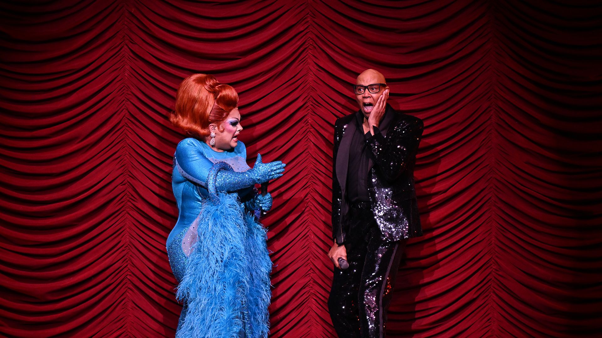 Two performers on stage in front of a red curtain. One in a sparkly blue dress with a feather boa and red hair, holding a microphone. The other in a black sequin suit, glasses, hand on face.