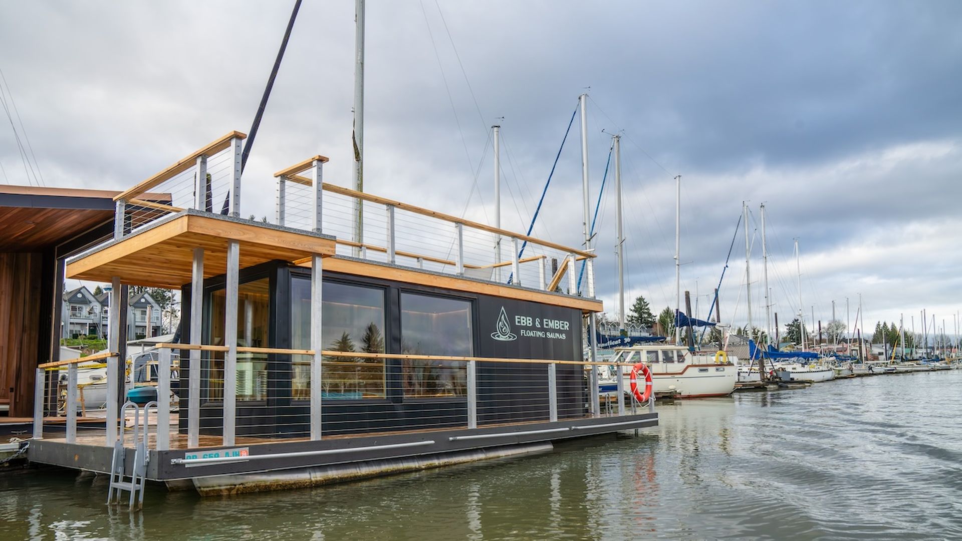 Black and wood floating sauna labeled "Ebb & Ember" docked on green water with sailboats and houses under a cloudy sky.