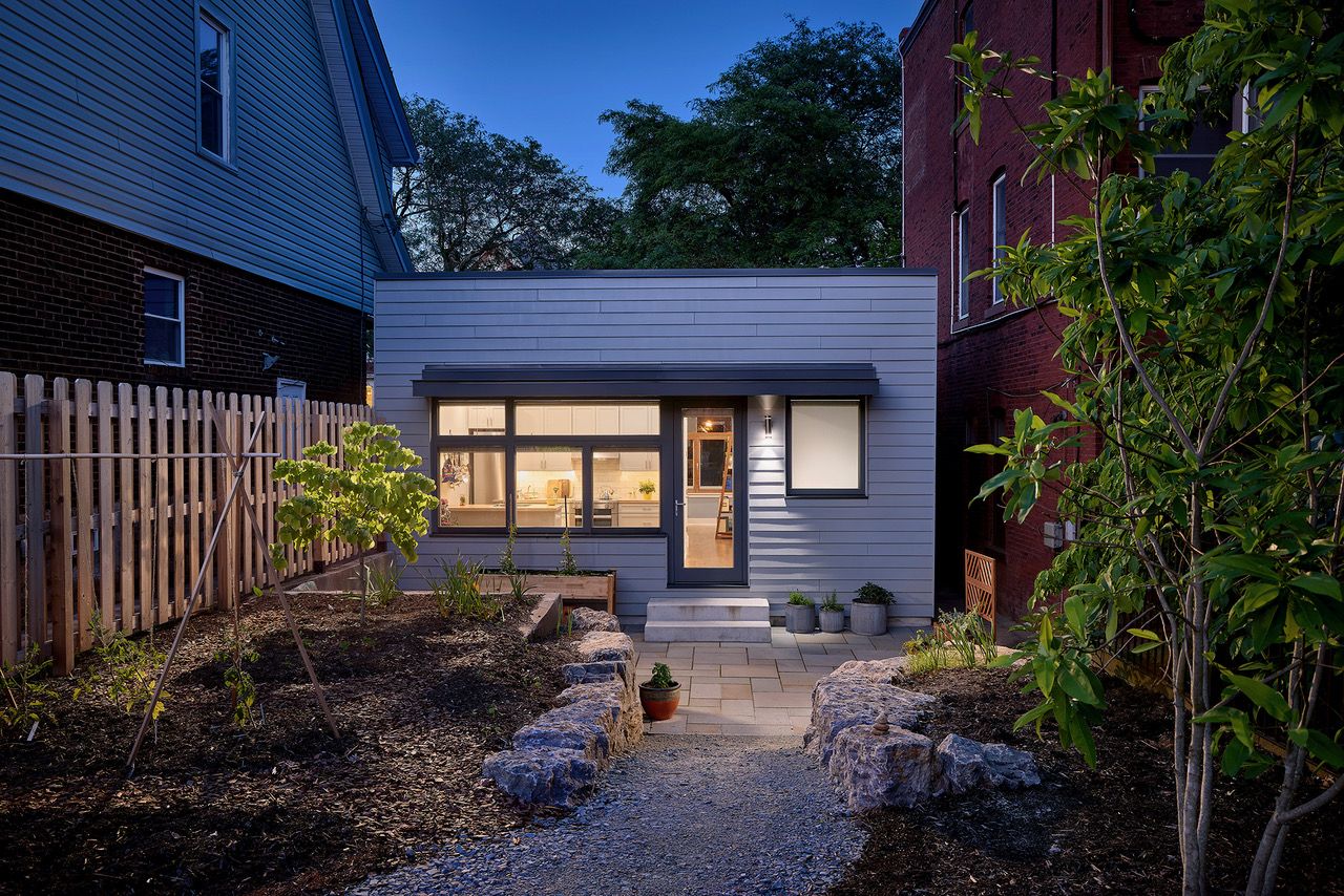 Modern single-story house with gray siding and large windows lit from inside, surrounded by garden beds with plants and stone edging, taken at dusk with a dark blue sky.