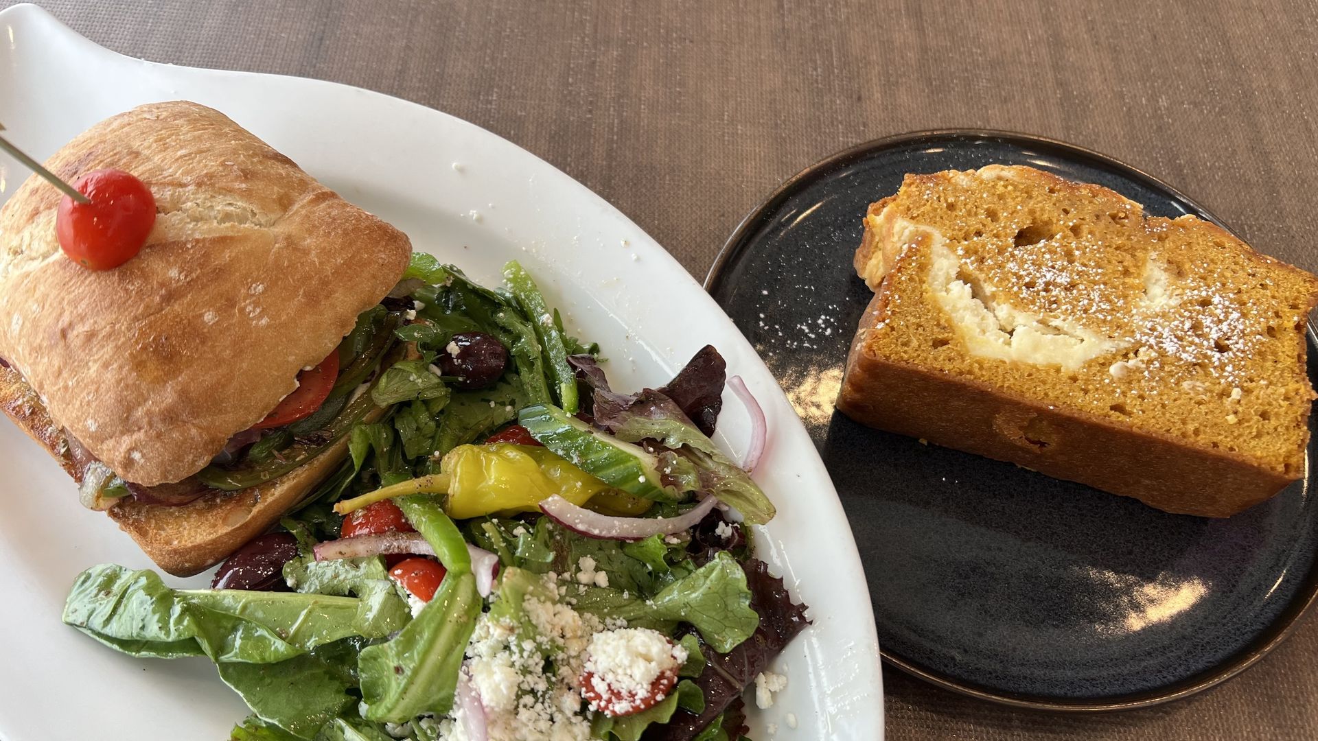 Sandwich with tomato and greens on white plate with mixed salad including pepperoncini, olives, red onions, cherry tomatoes, and cheese, next to a slice of ound cake on a dark blue plate.