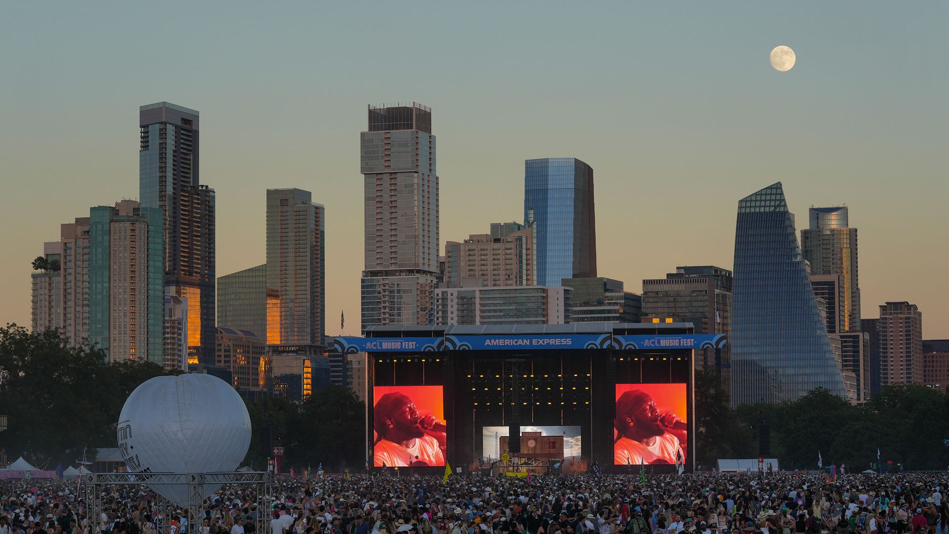 City skyline at sunset with a huge outdoor concert crowd before glassy towers. Stage banner reads "ACL Music Fest"; a large white balloon on the left and a bright full moon in the sky.