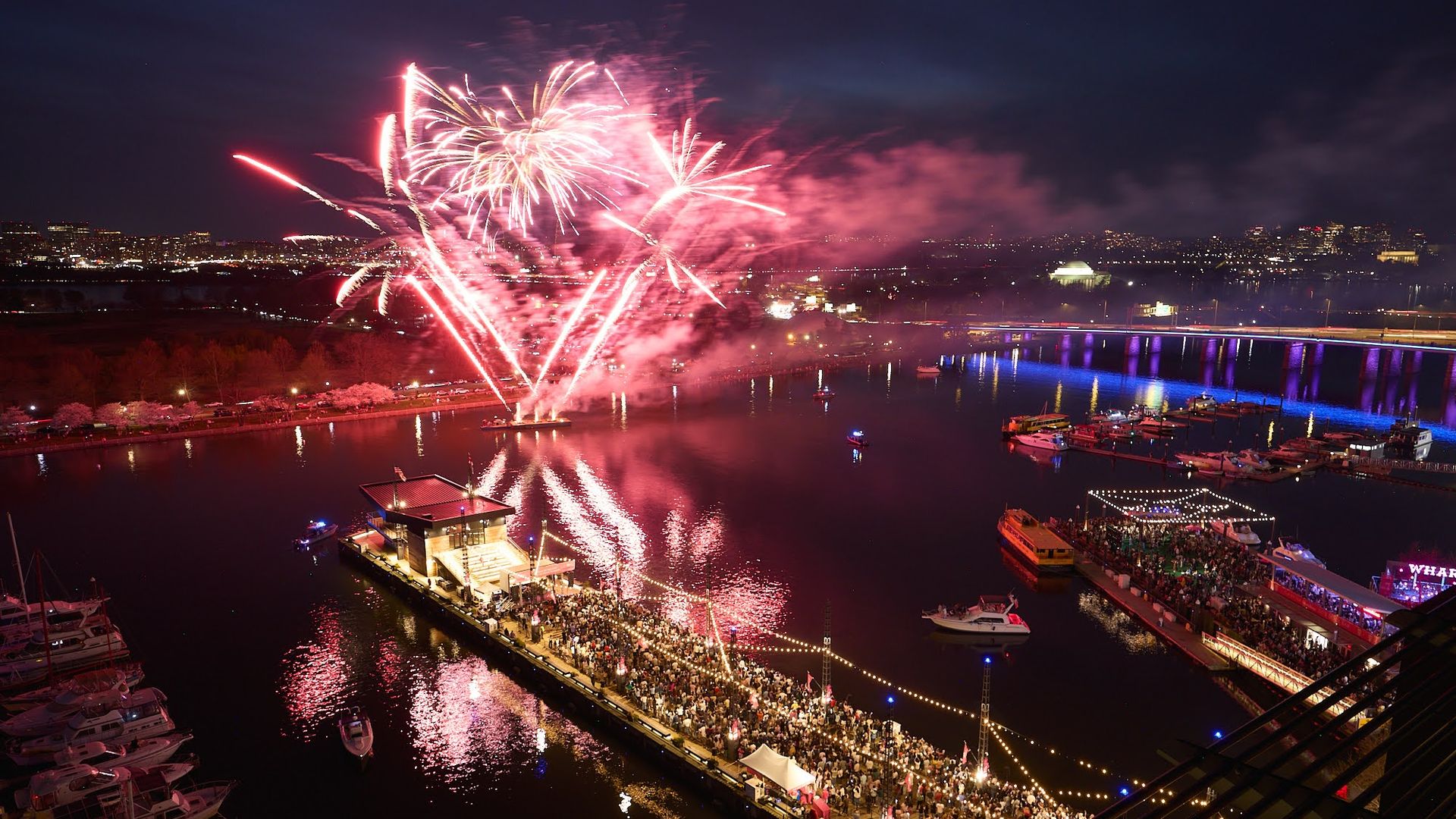 Nighttime harbor with pink fireworks over a floating stage and crowded pier. Reflections glow on calm water; boats docked at the marina, blue-lit bridge in the background and city lights.