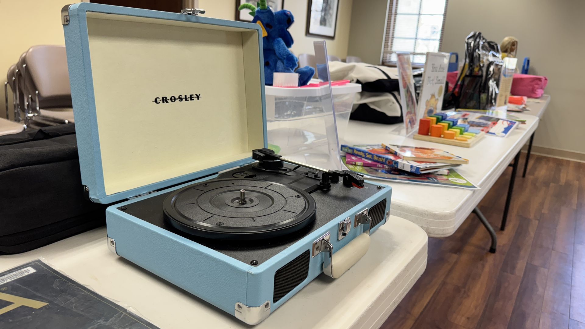 An open blue Crosley record player sits on a table in a bright room, with colorful toys and a blue plush dragon nearby; framed pictures on the wall and a window with blinds in the background.