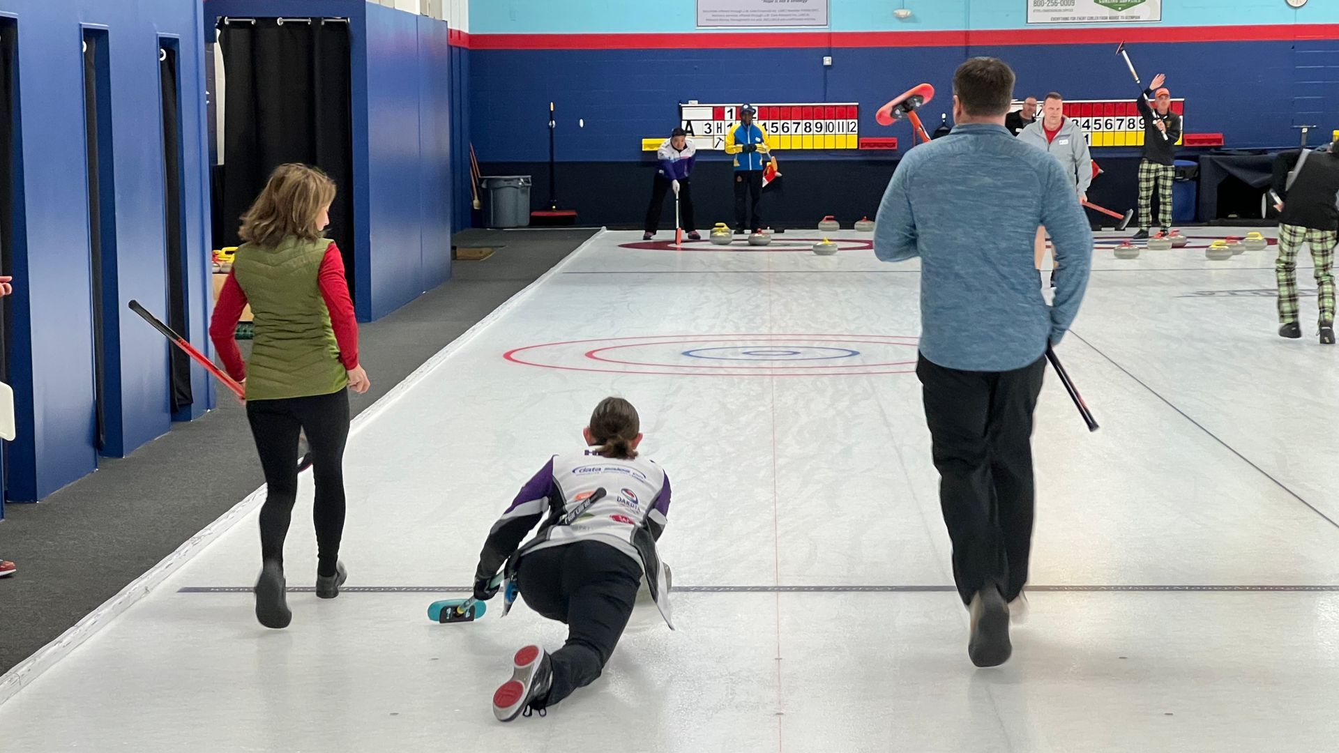 A curler throws a stone down a curling rink towards a target located away from the camera, with two sweepers running on either side of her.