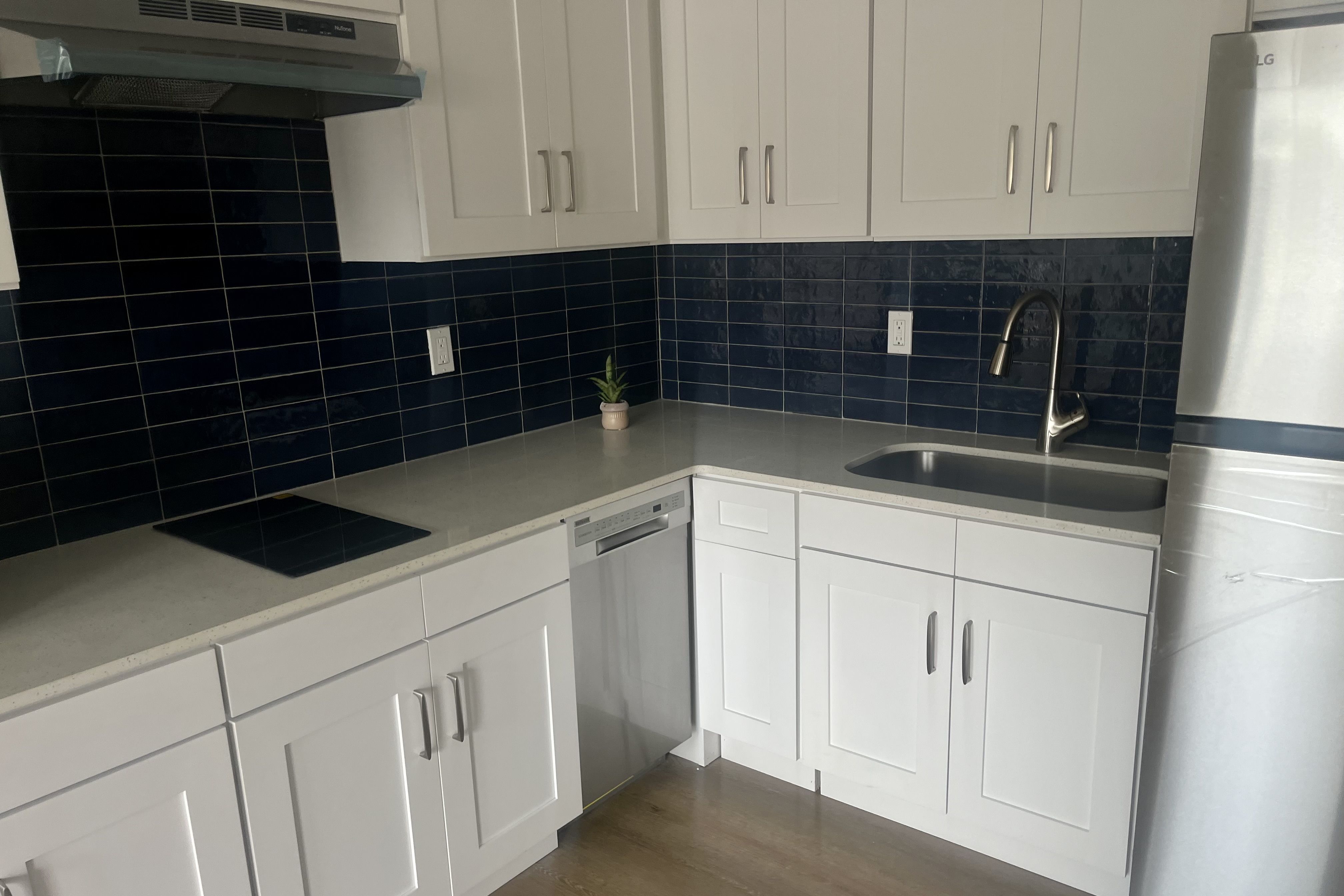 Modern kitchen corner with white cabinets, light gray countertops, dark blue rectangular tile backsplash, stainless steel sink, faucet, dishwasher, and a refrigerator on wooden floor.
