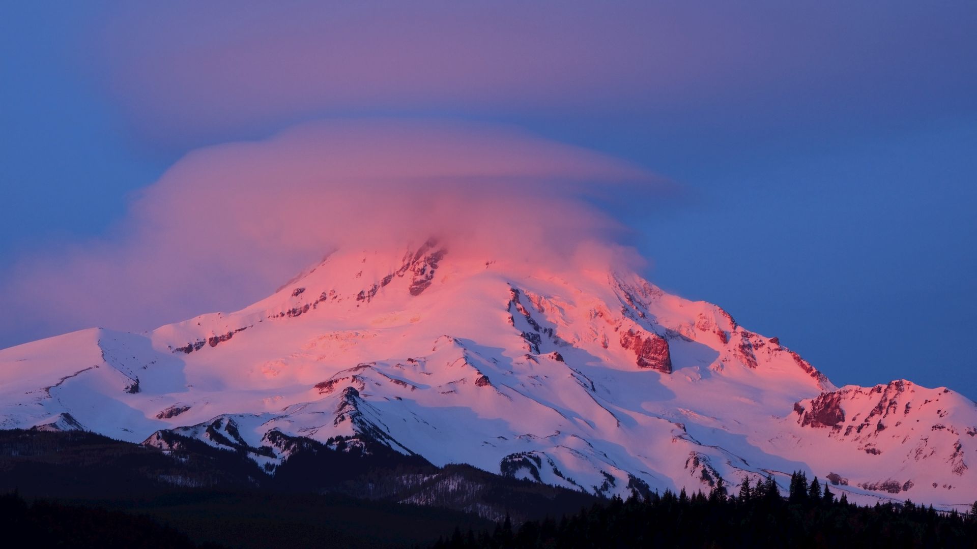 Dramatic pink clouds drape the top of a snow covered Mount Hood at sunset.
