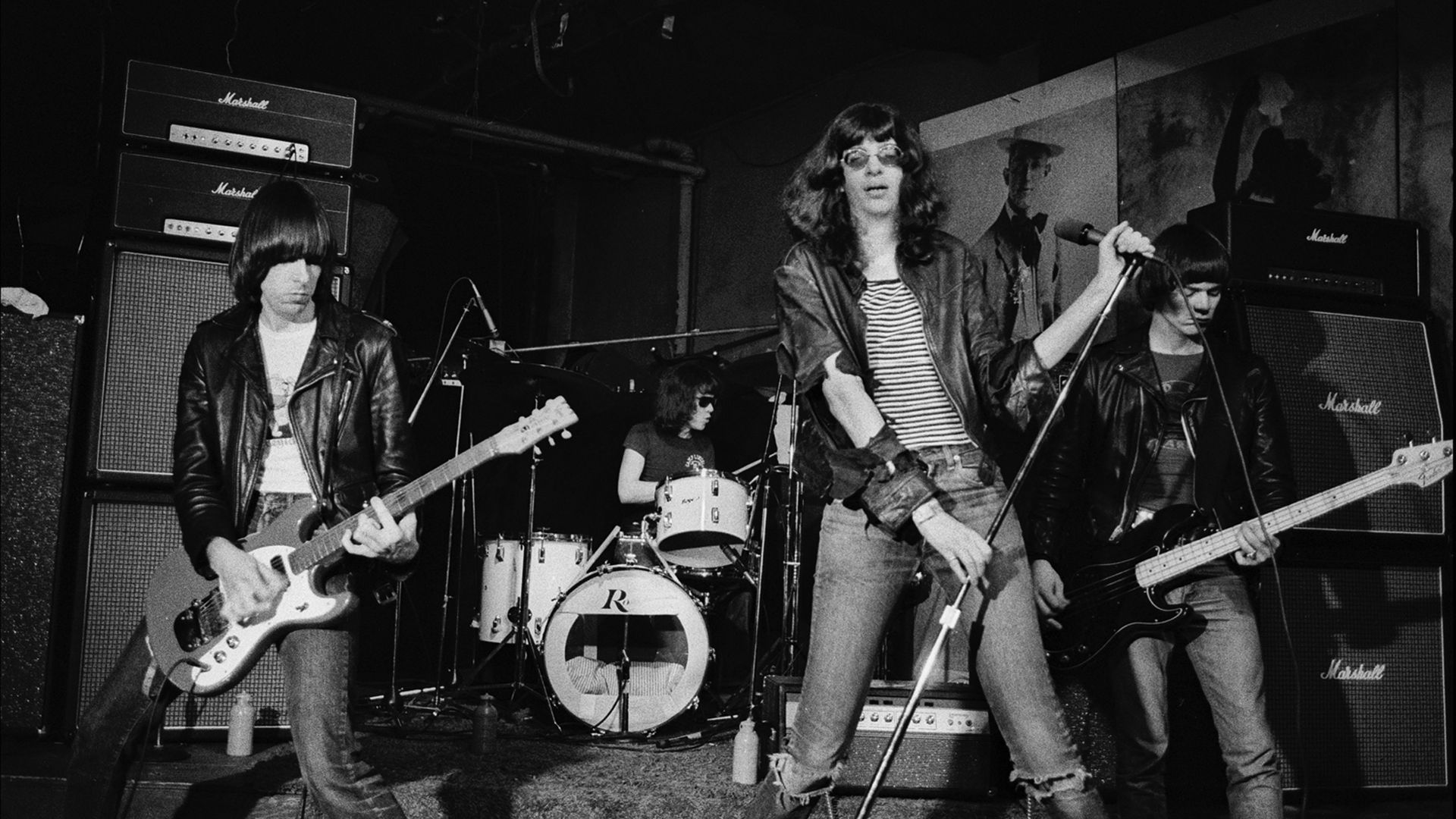 Black-and-white photo of a rock band on a small stage: lead singer in striped shirt and leather jacket center, guitarist left, bassist right, drummer behind, Marshall amps visible.