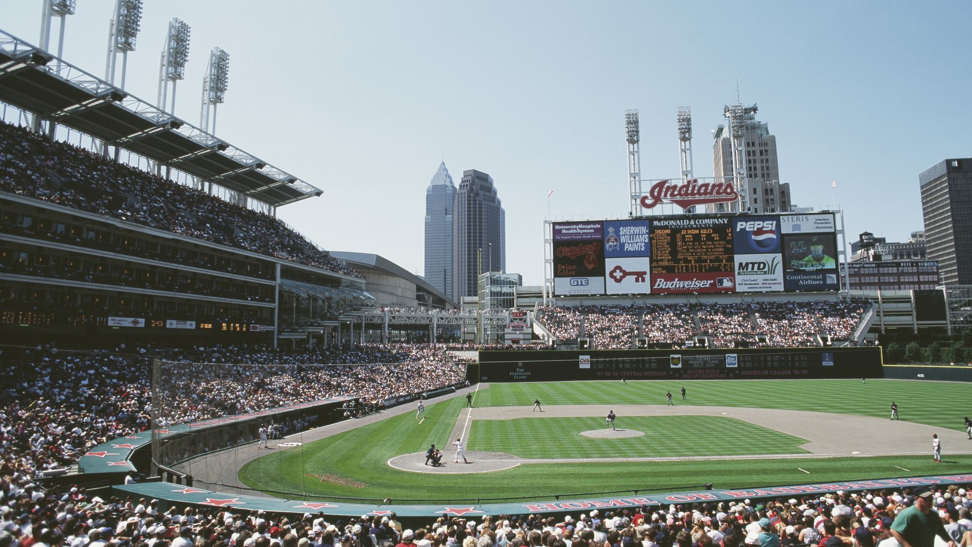 Wide view of a crowded baseball stadium, green field, and players; a large scoreboard reads "Indians" with ads, while a city skyline rises in the background under a clear blue sky.