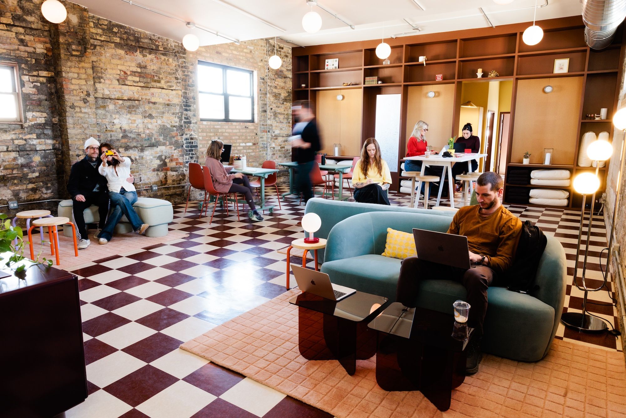 Bright coworking space with exposed brick walls, a black-and-white checkered floor, and round pendant lights. People work at tables on laptops or relax on couches, with tall wood shelving along the back wall.