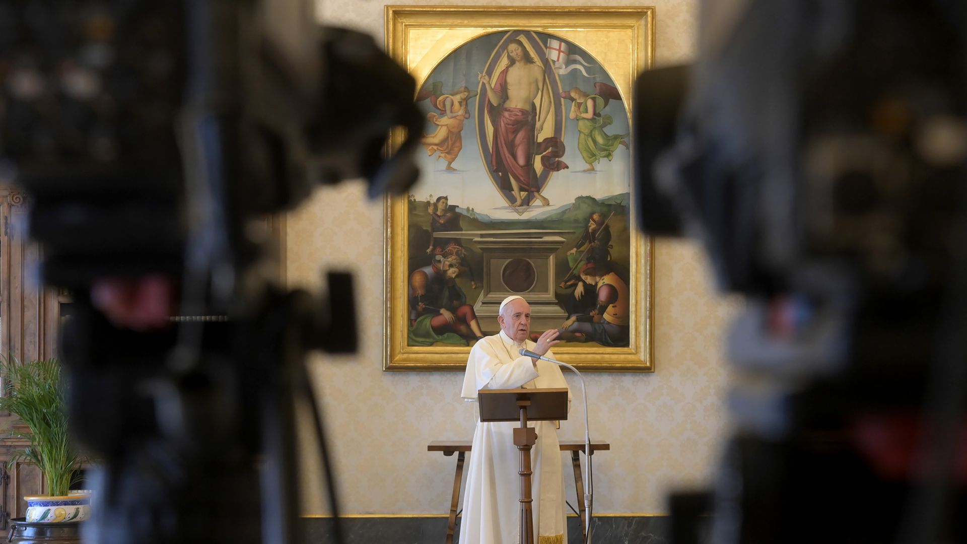In this image, Pope Francis stands and speaks in front of two cameras.