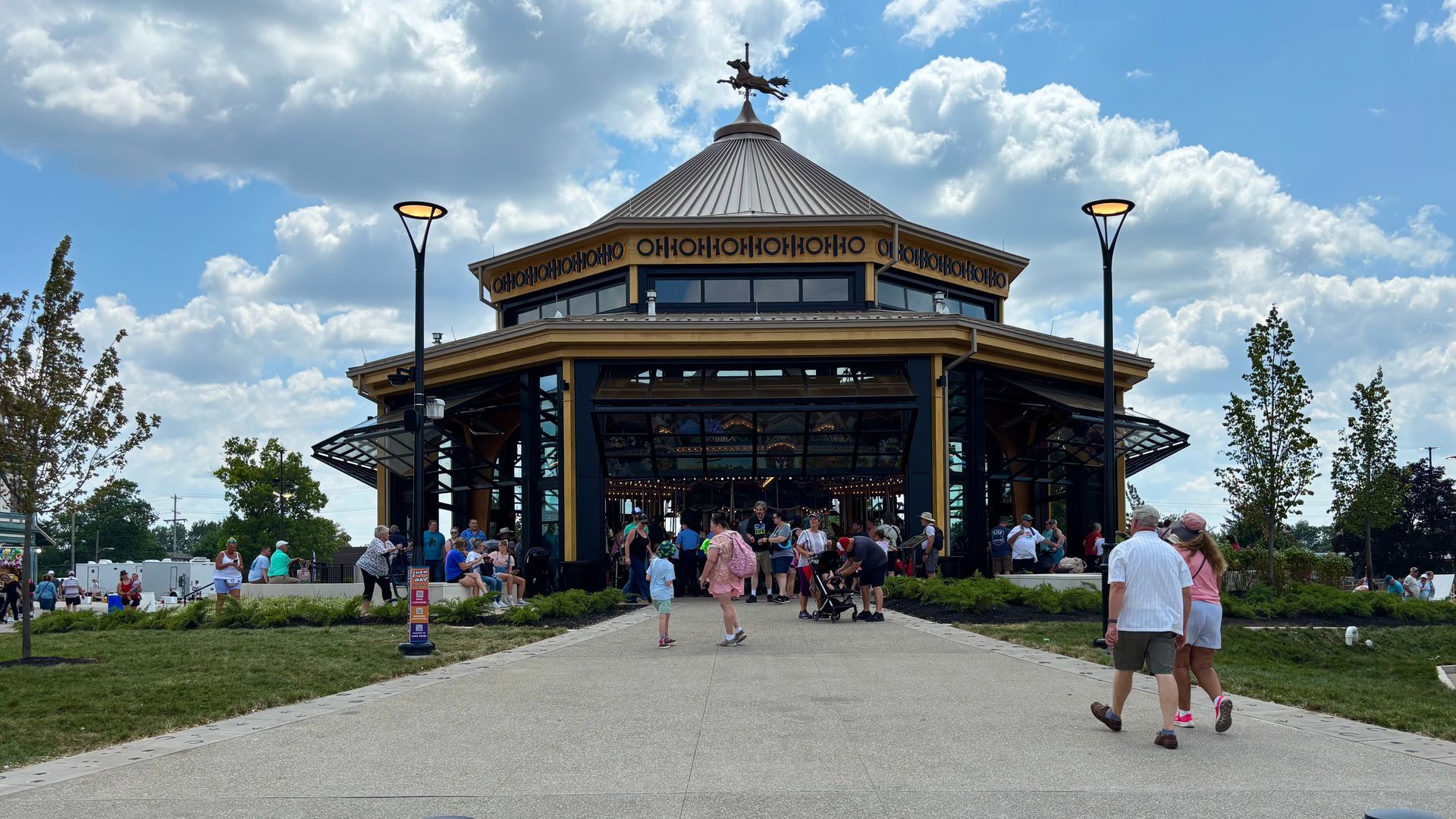 Crowds gather outside a glass-front carousel pavilion with a tall conical metal roof and statue on top, beneath a blue sky with white clouds.