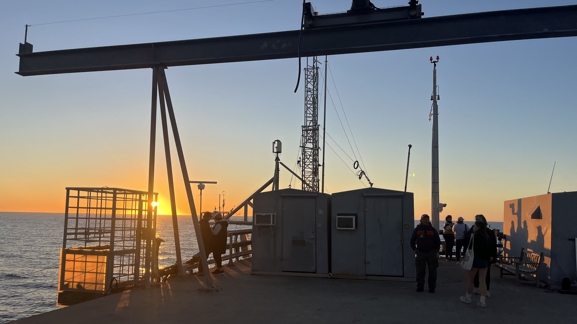 Sunset view over the ocean from a pier with silhouetted people, metal structures, and two small gray buildings on a concrete platform.