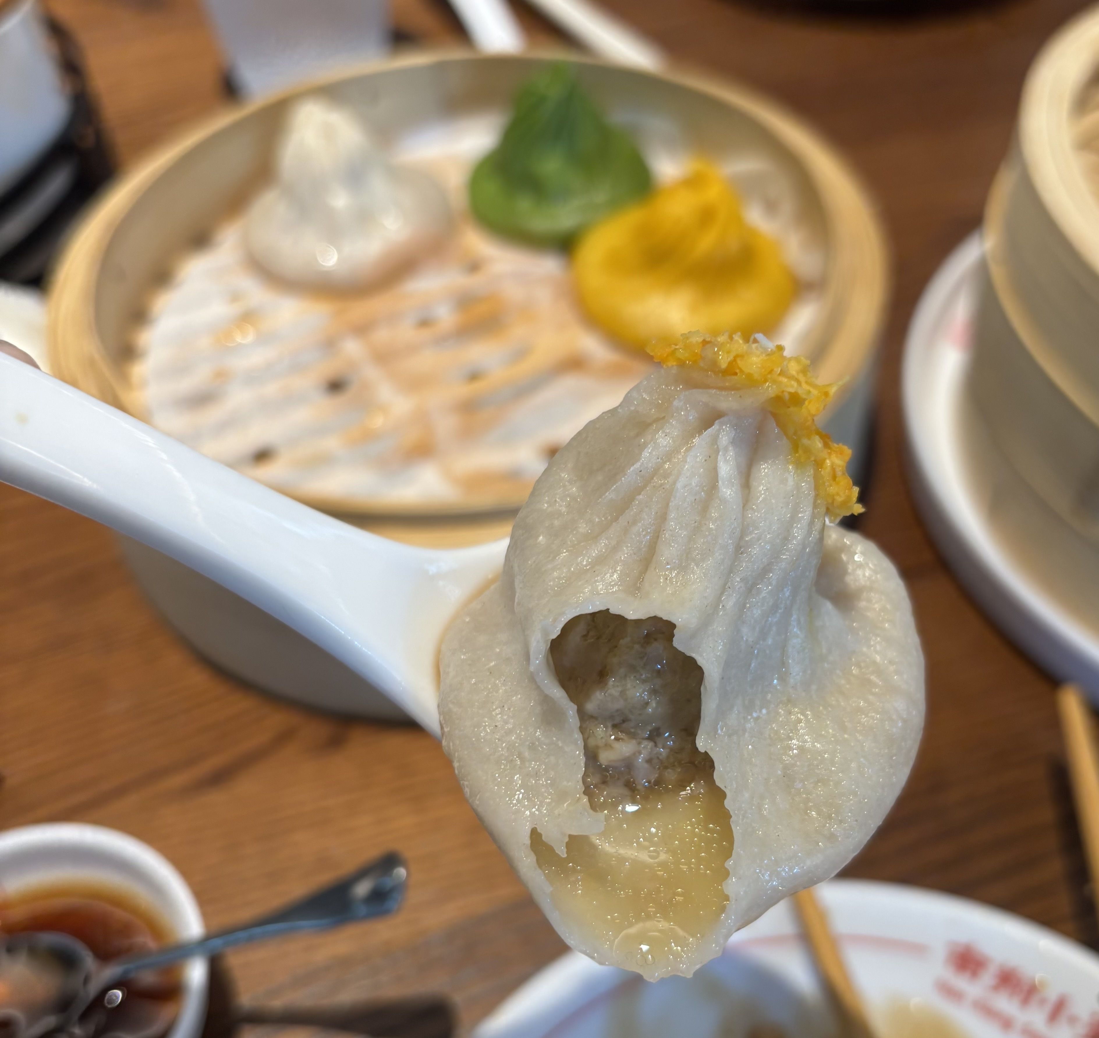 Steamed dumpling on a white spoon, bitten open to show broth inside and a crispy orange topping; background shows a bamboo steamer with more dumplings.