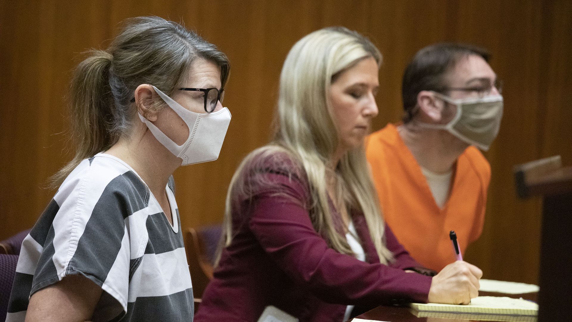 Jennifer Crumbley on the left in a striped gray/white shirt wearing a mask, attorney in the middle, and James Crumbley on the left in an orange shirt and mask. 
