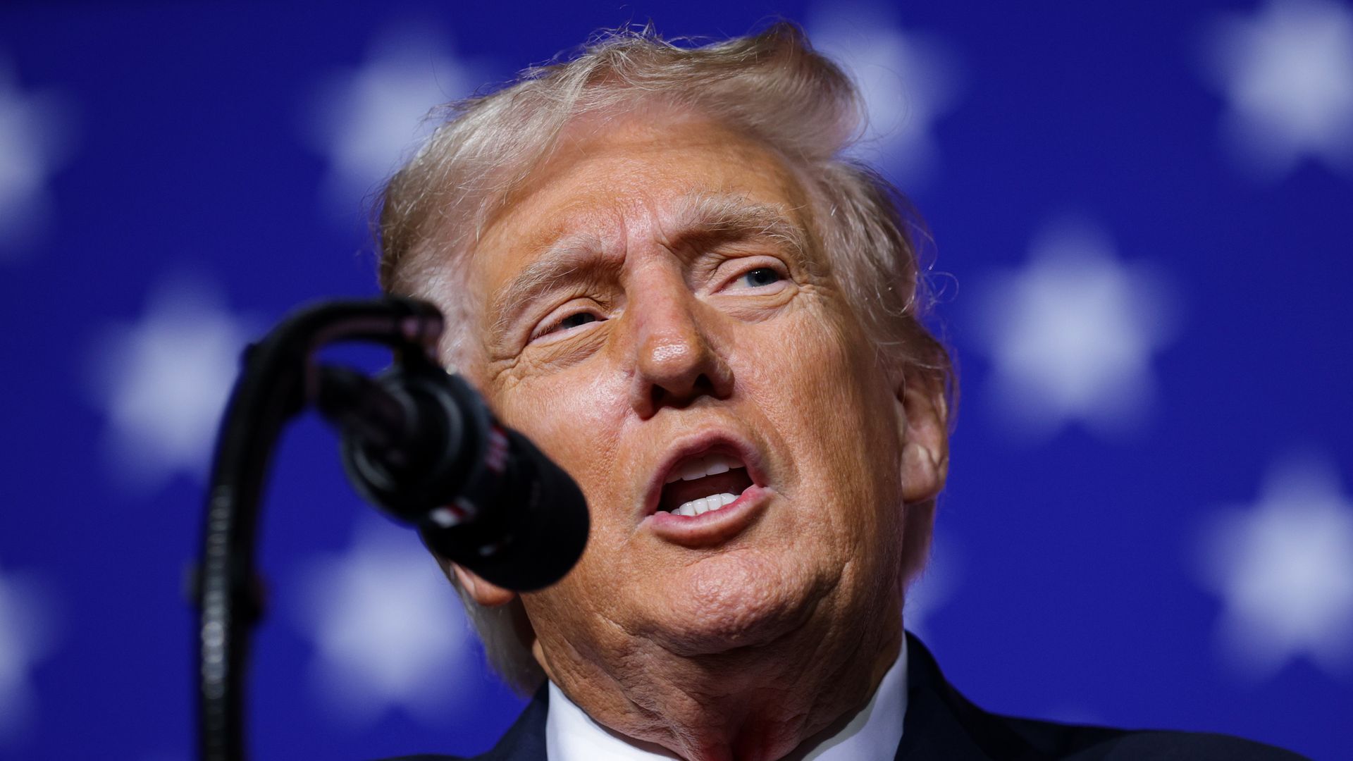 President Trump, wearing a navy jacket with a US flag, white shirt and red tie, speaks into a black mic in front of the white stars and blue background part of a large US flag.