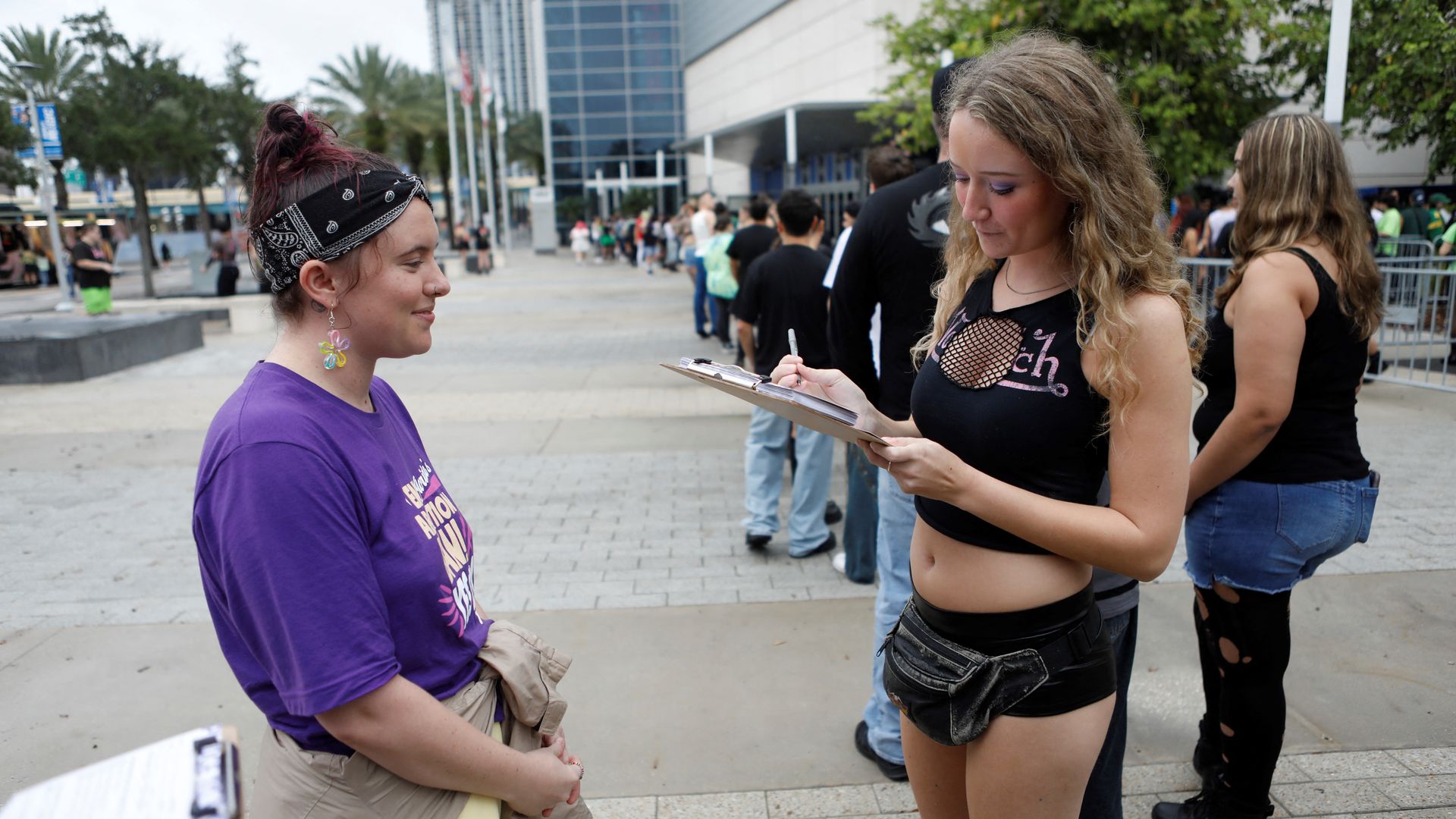 A person wearing a purple T-shirt and black bandana smiles at a person in an all-black outfit looking down at a clipboard.
