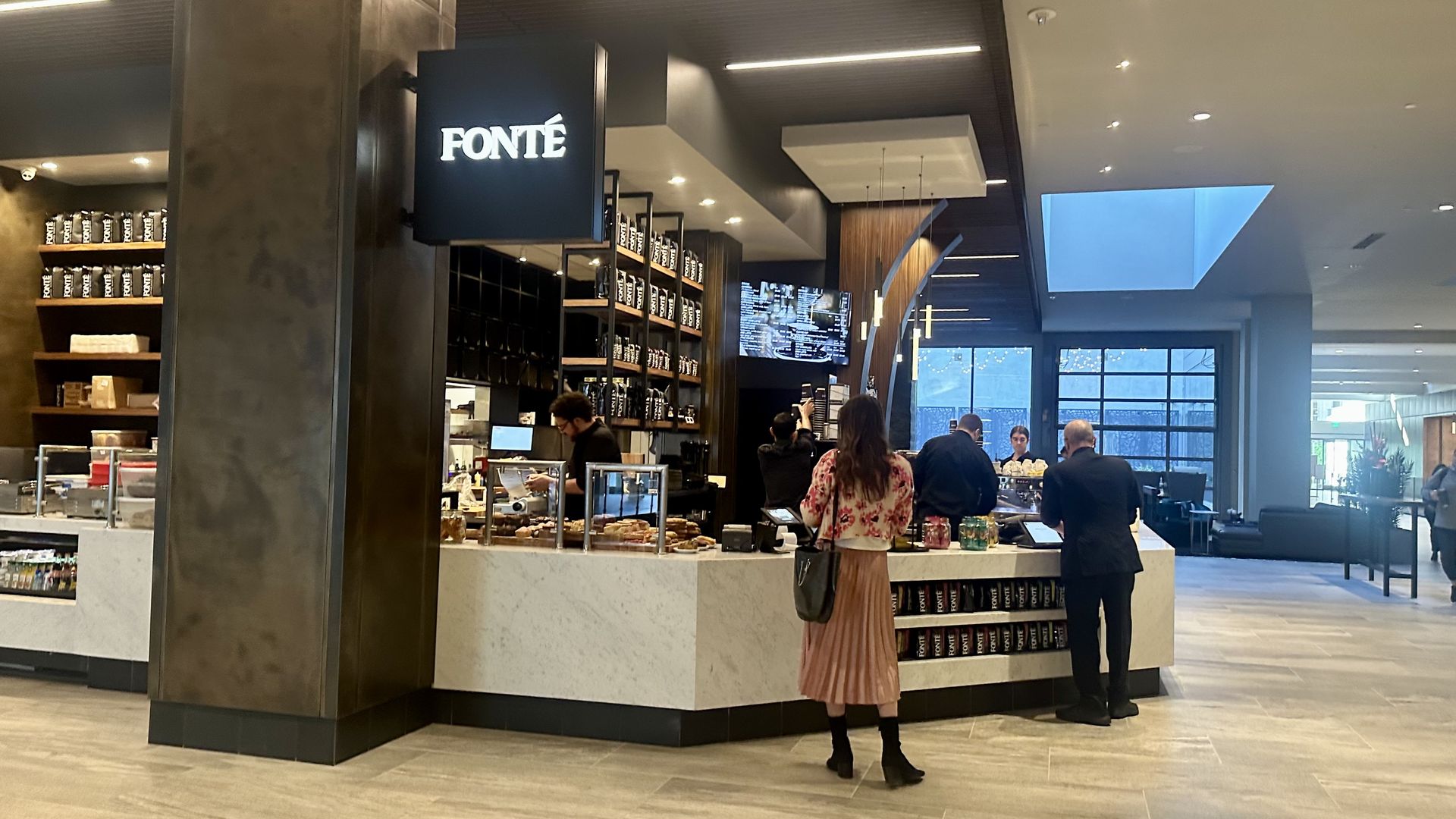 Two people stand near a light colored coffee counter with dark wood accents behind and a sign that says "Fonte"