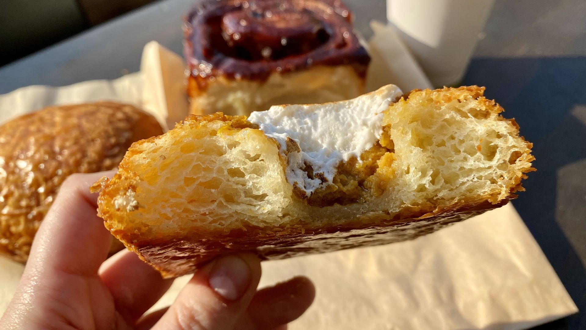 A hand holds a croissant that is bitten into already, with orange colored filling and a smear of white on top, with other pastries and a  cappuccino in background.