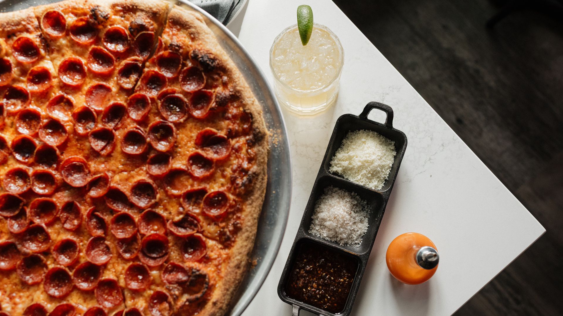 Pepperoni pizza on a metal tray with a textured crust, next to a three-section container holding grated cheese, coarse salt, and chili flakes, a small hot sauce bottle, and a lime garnish drink on white table.