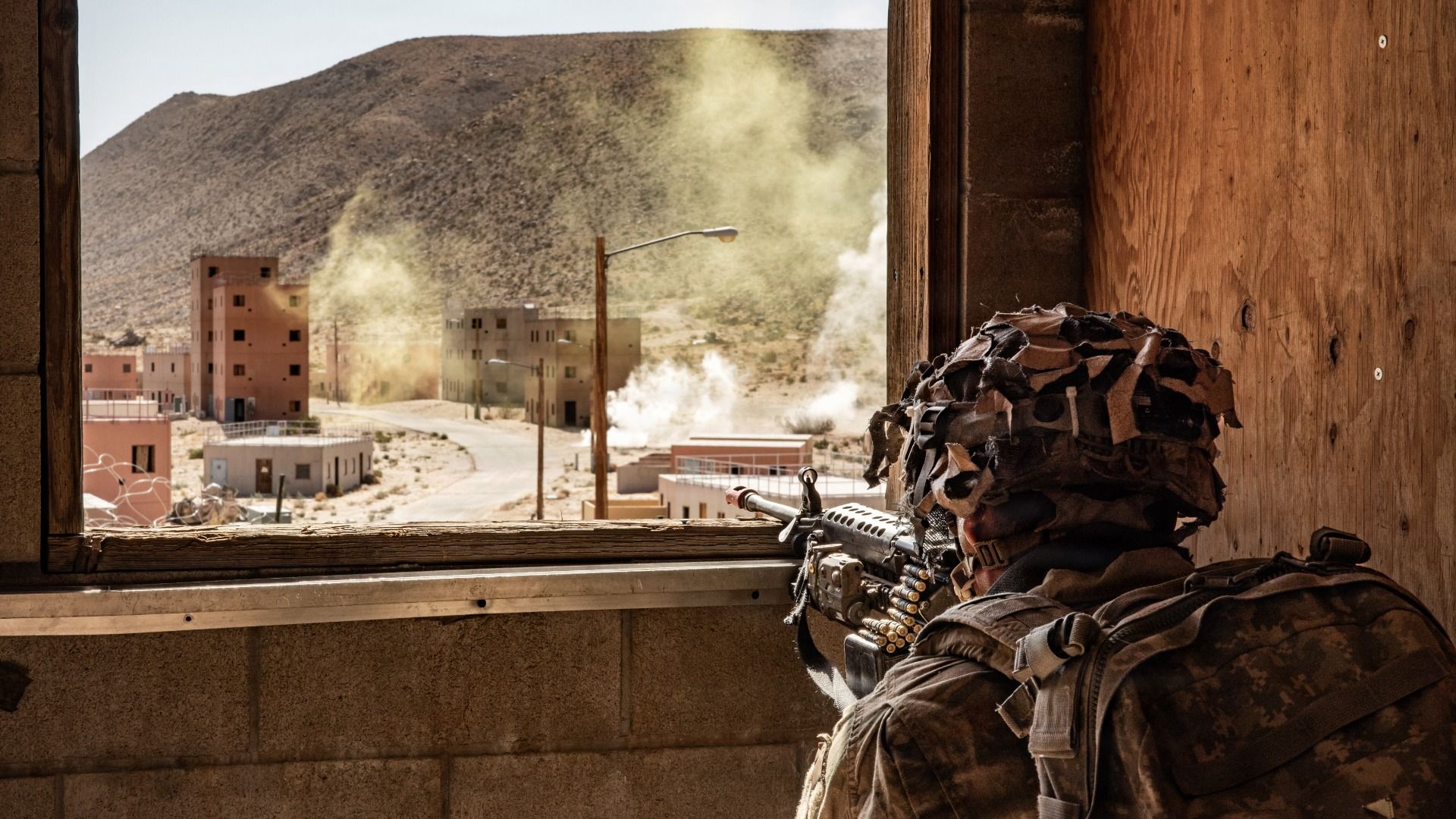 An American soldier in camouflage, peering from a wood-framed window with a rifle and ammo belt, overlooking a dusty town with smoke plumes, beige buildings, and a rocky hill.