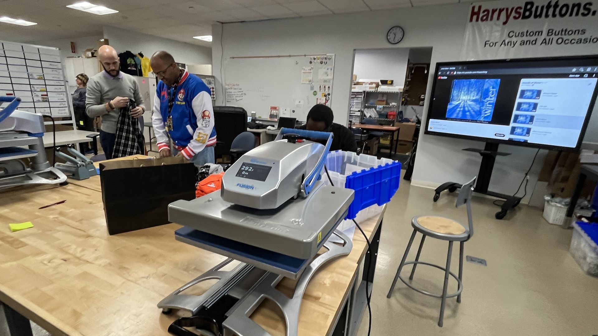 Interior of a custom button workshop with three men working. Large heat press machine on wooden table in foreground. Screen on wall shows winter-themed video. Sign reads HarrysButtons.