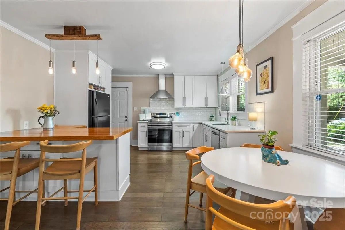 Bright kitchen with white cabinets, stainless steel appliances, wooden floor, a wooden breakfast bar with three chairs, a white round dining table with four chairs, and pendant lights.