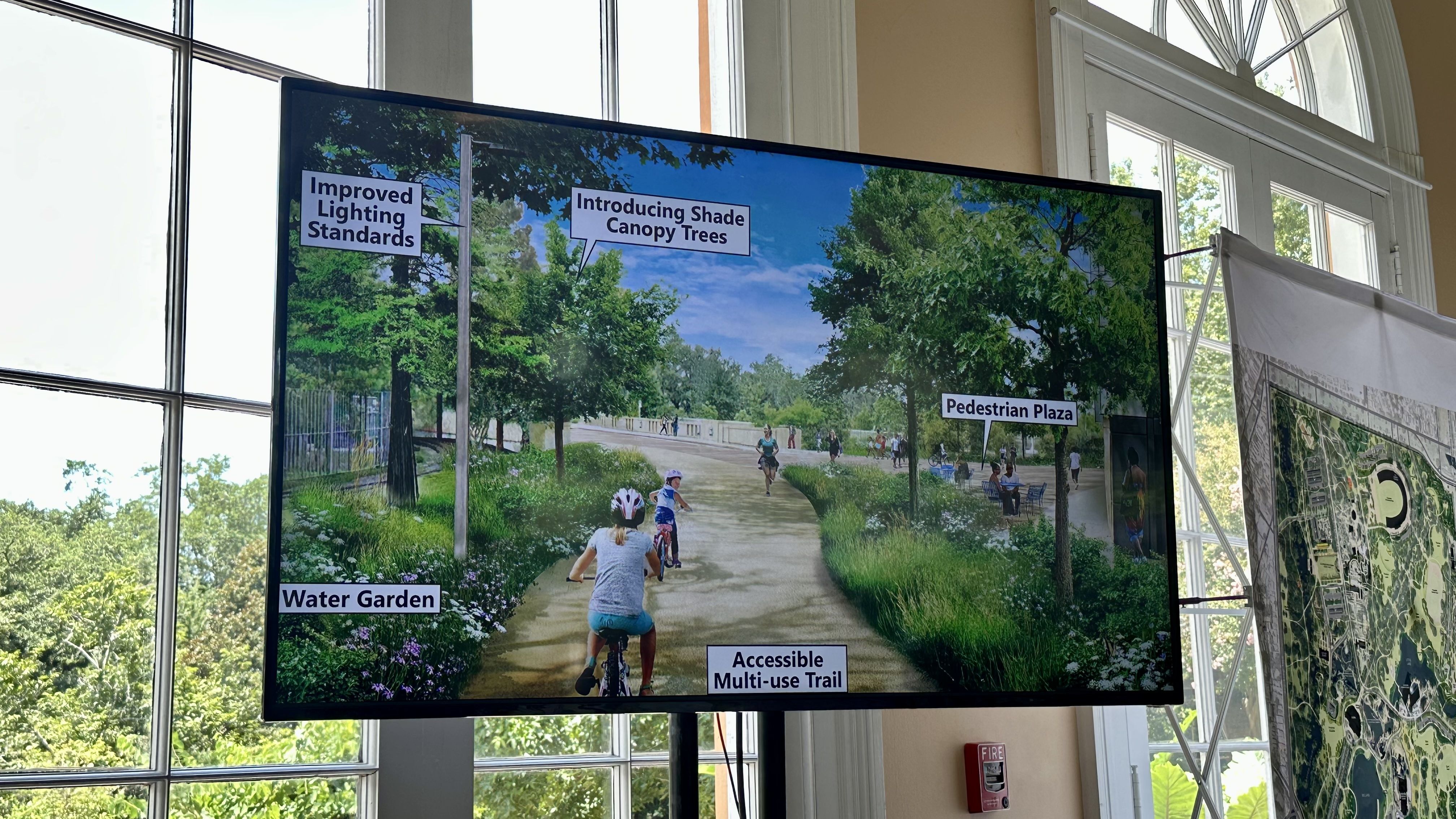 The image shows a digital rendering displayed on a screen in a bright room with large windows. The rendering depicts a multi-use trail in a park with various enhancements labeled: 'Improved Lighting Standards,' 'Introducing Shade Canopy Trees,' 'Water Garden,' 'Accessible Multi-use Trail.