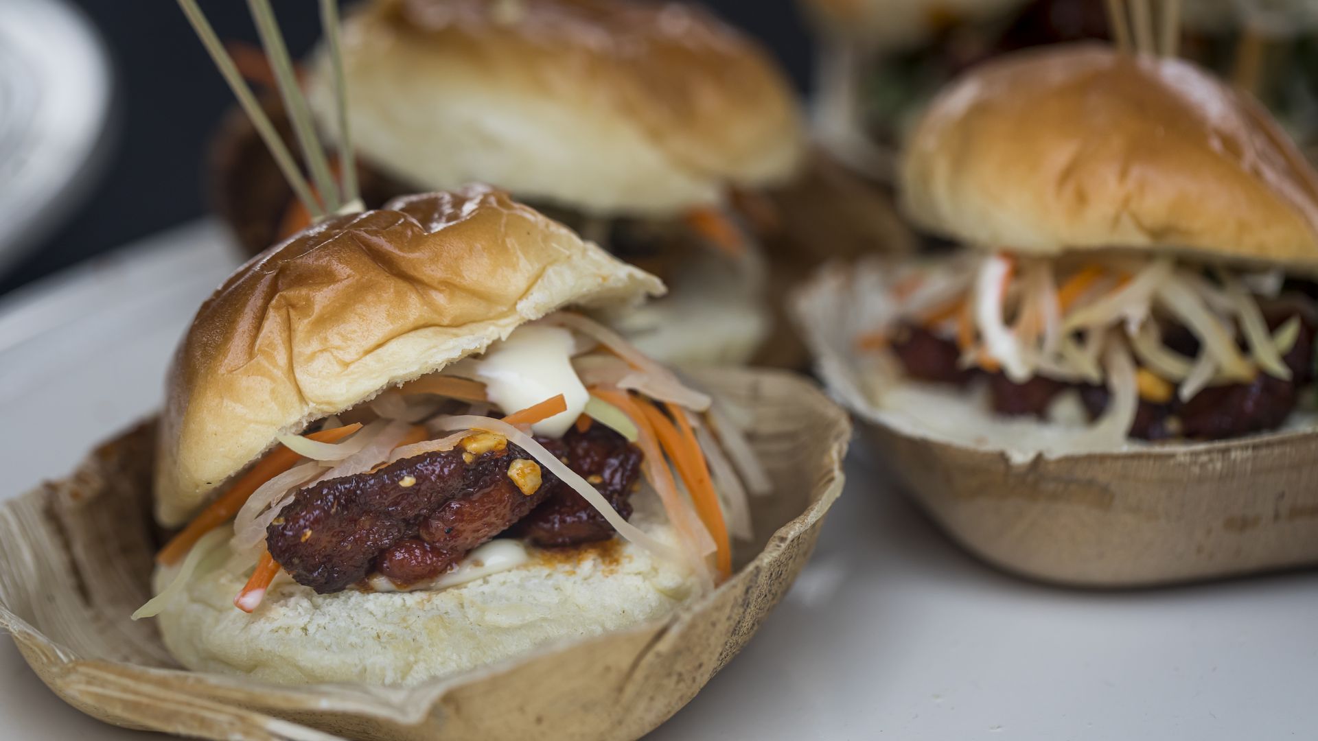 Close-up of small sandwiches with glossy golden buns, filled with dark grilled meat, shredded white and orange vegetables, and a creamy white sauce, served in brown plastic bowls.