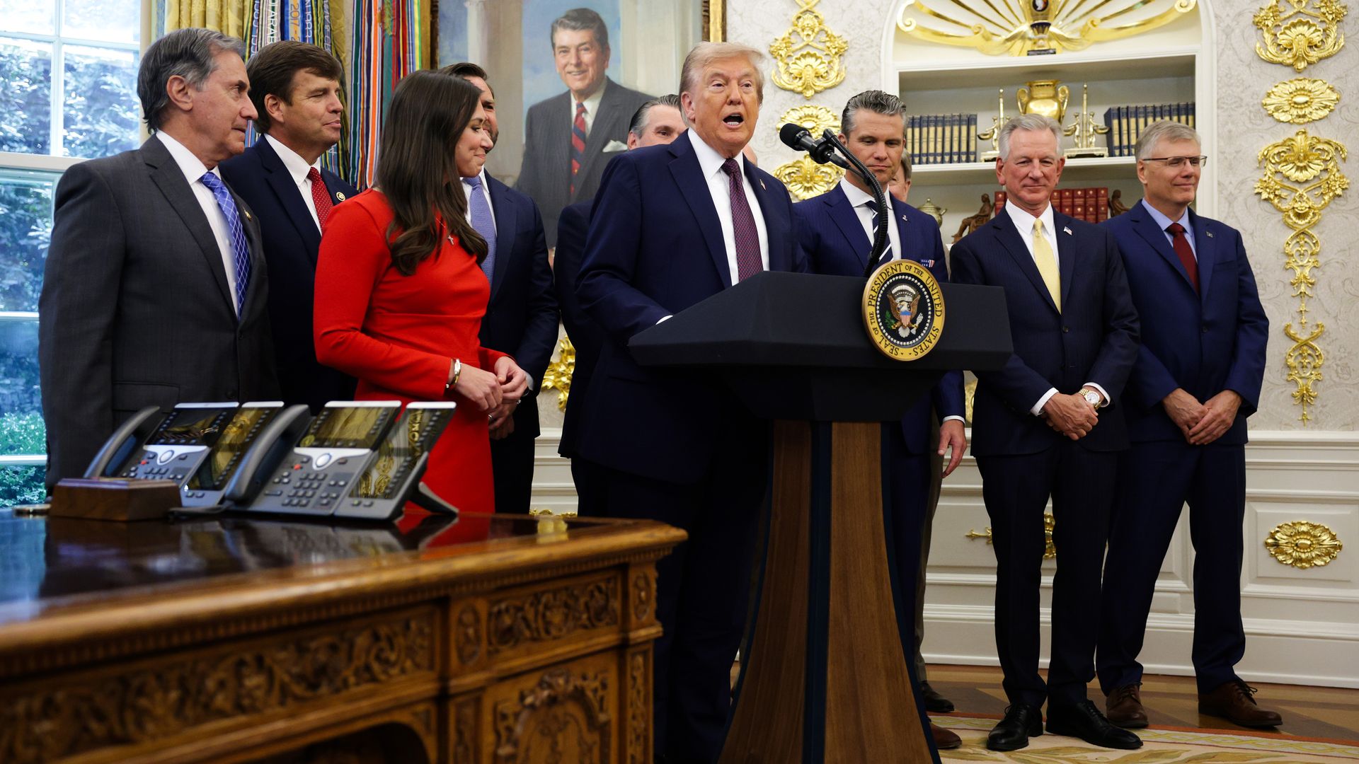 Lawmakers and President Trump gather in the Oval Office. Trump is speaking behind a podium. A window and wall decorations can be seen.