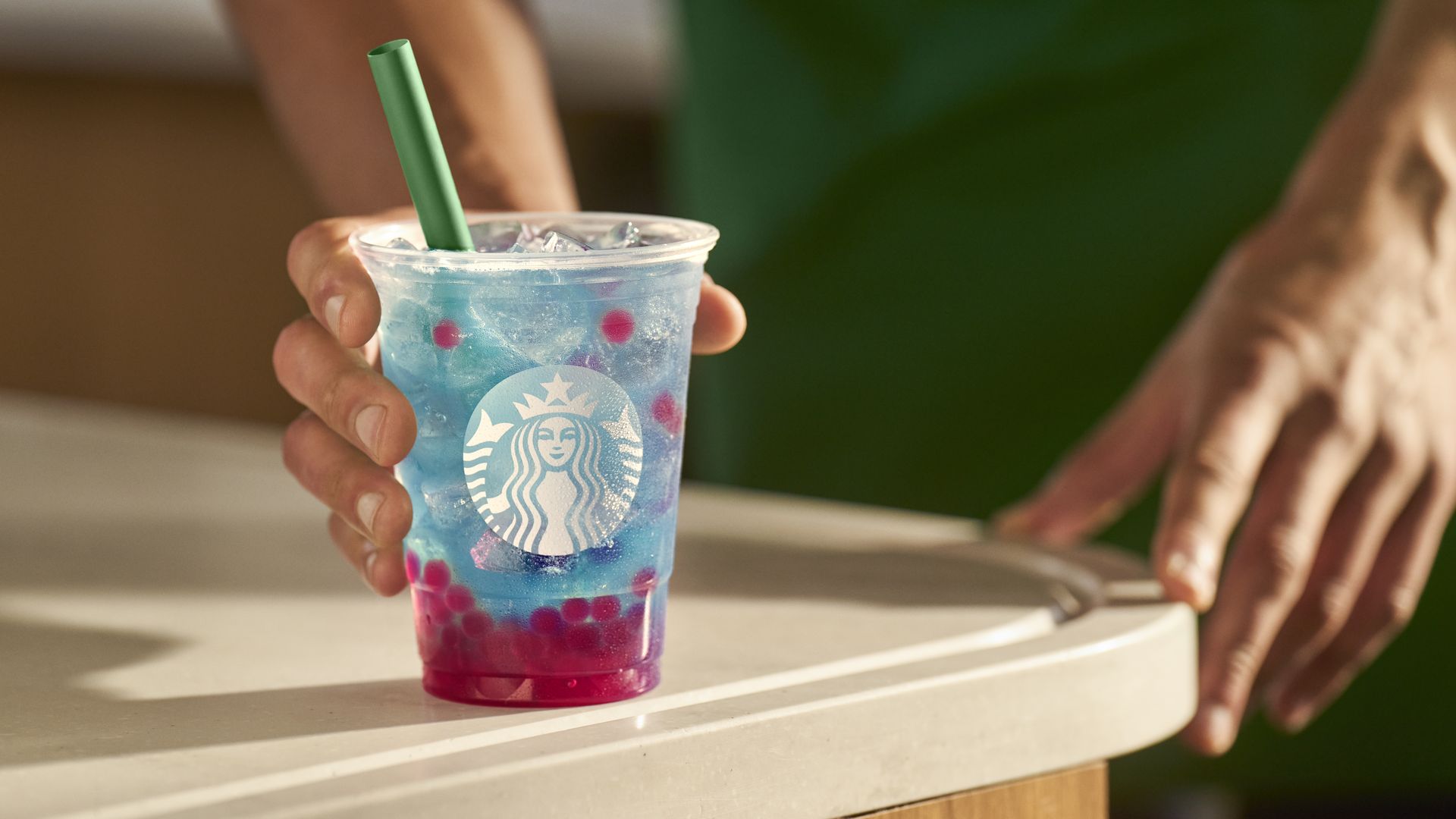 A Starbucks barista wearing a green apron holds a Summer-Berry Refresher, a blue drink with pink berries in a clear cup. The cup has a green straw in it. 