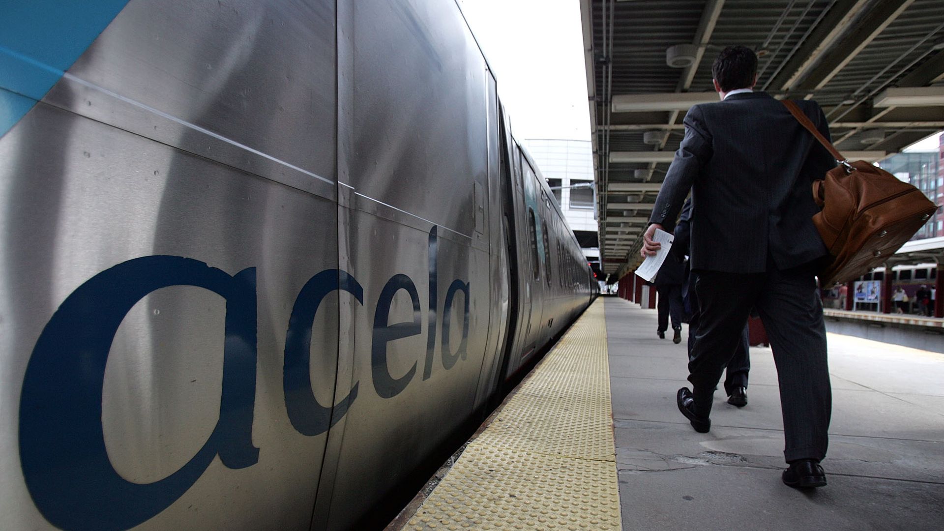 People in business attire walk on a platform beside a silver Acela train with blue lettering at a train station under a roofed structure.