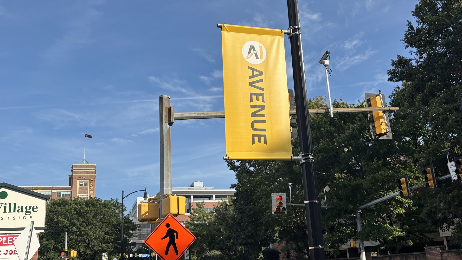 Bright yellow vertical street banner reading AVENUE with a circular logo, attached to a black lamppost; orange diamond construction sign with a worker icon in the foreground, blue sky, trees, and city buildings in background.