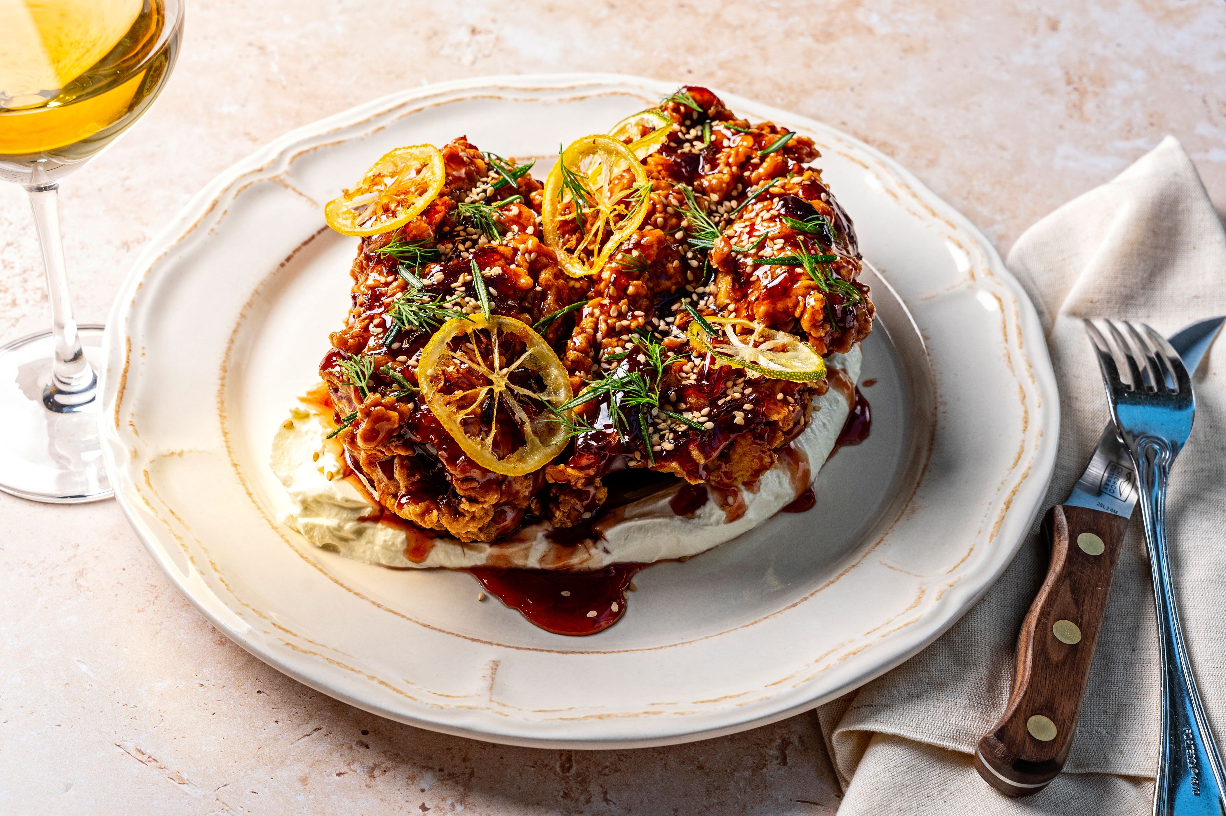 Two pieces of crispy fried chicken glazed in dark sauce, topped with sesame seeds, lemon wheels, and dill sprigs, served on a white plate with a beige napkin, alongside a wine glass and cutlery.