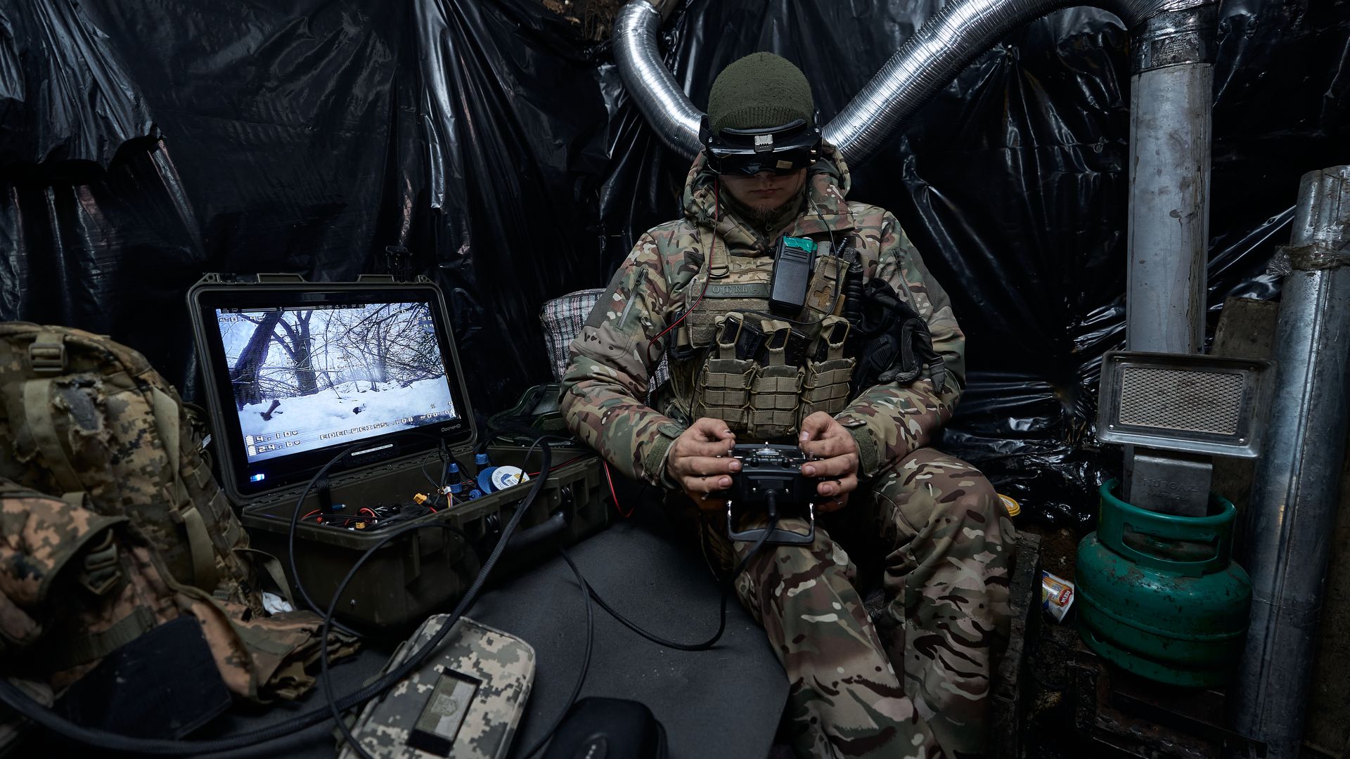 A member of the Ukrainian military sits in a blacked-out room. He is wearing a headset and manipulating a controller. A screen is illuminated to his right.