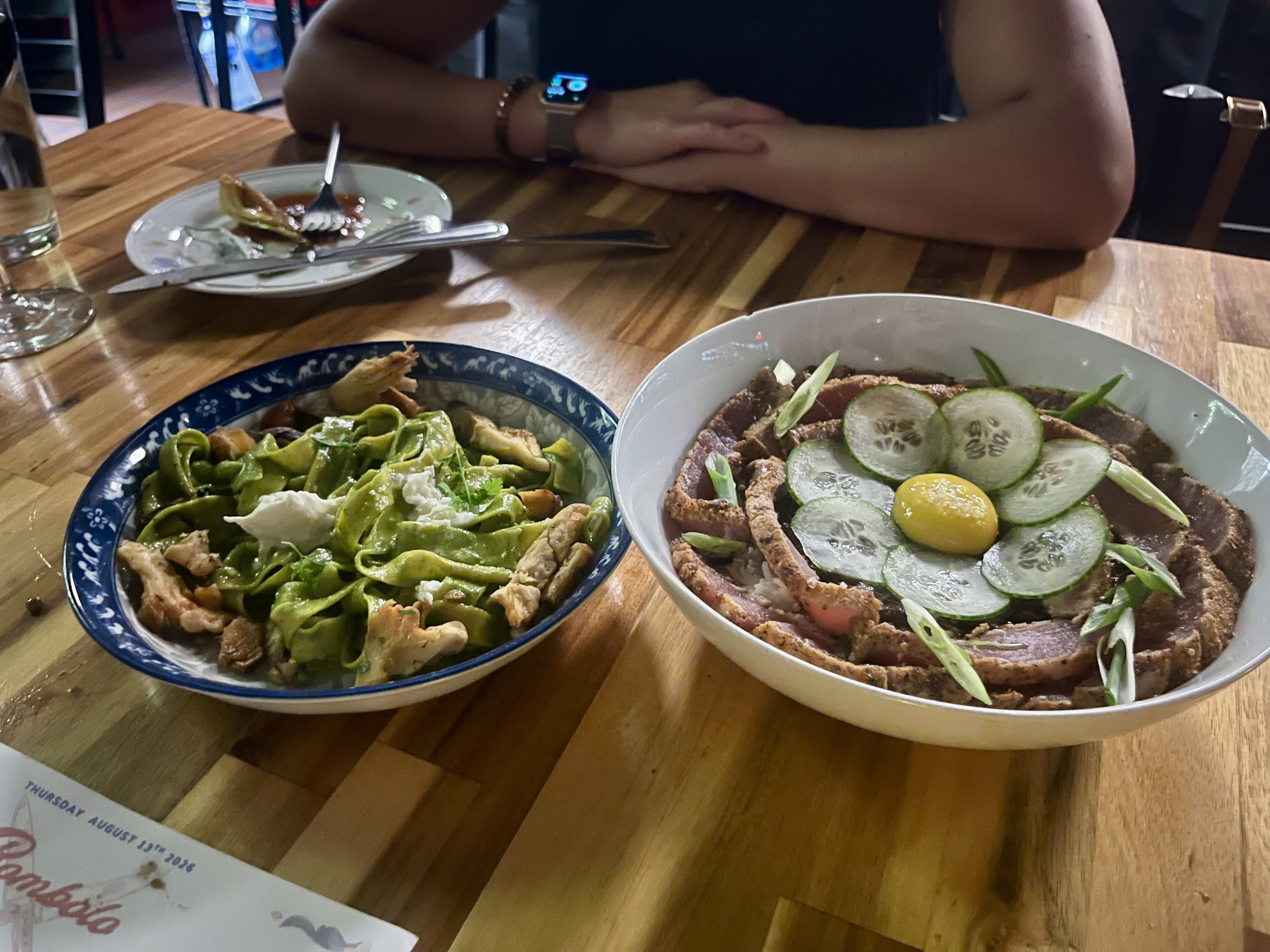 Person sits with arms folded at a wooden table. Two dishes: a blue-rimmed bowl of green pasta with chicken, and a white bowl topped with cucumber slices, greens, and a yellow egg yolk.