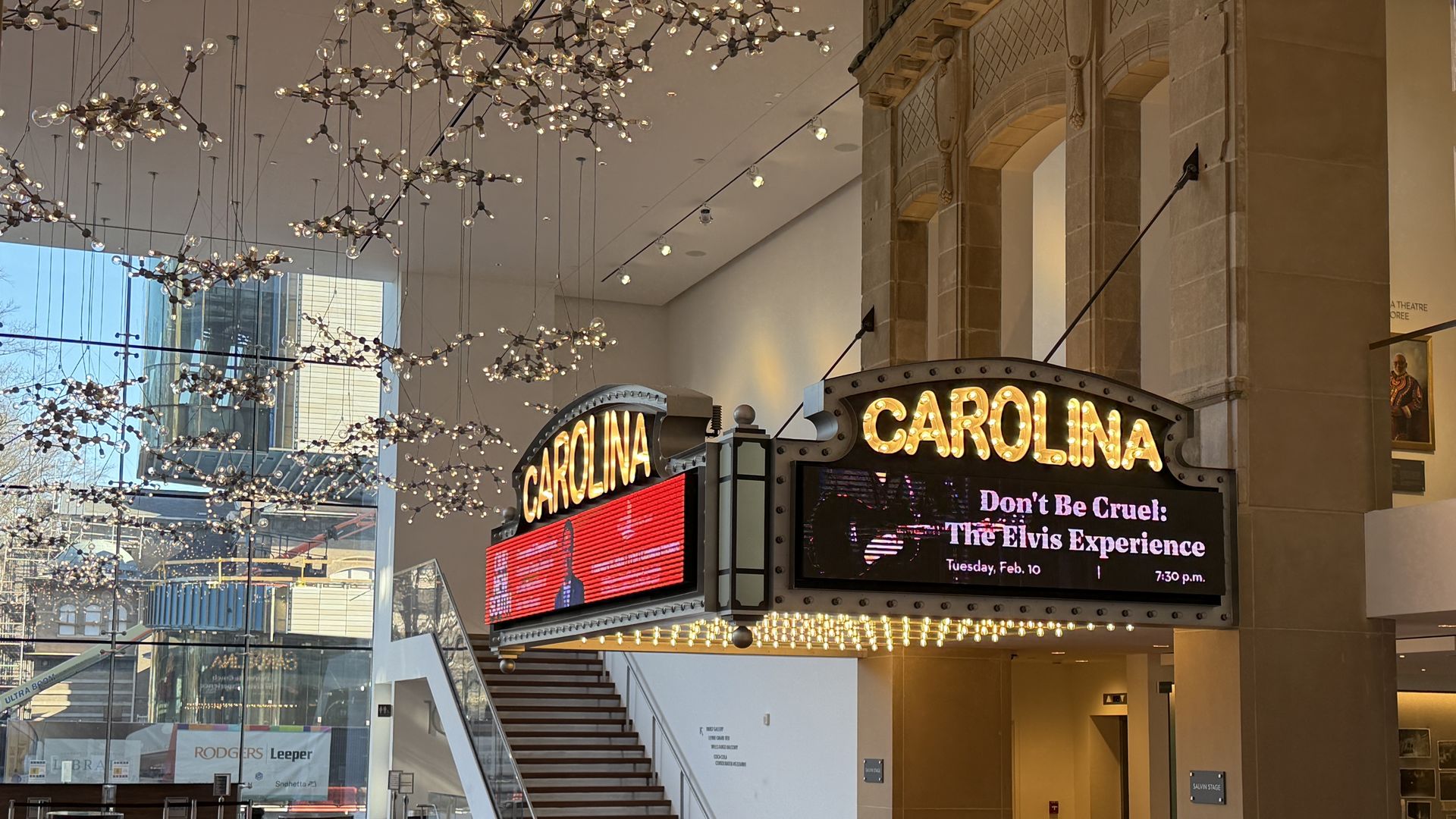Lobby with wooden floor and modern hanging lights, featuring an illuminated theater marquee reading "CAROLINA" and advertising "Don't Be Cruel: The Elvis Experience" for Feb 10 at 7:30 p.m.