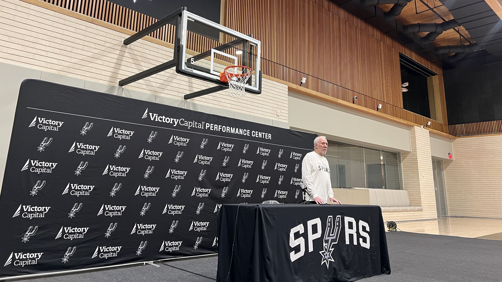 Gregg Popovich stands at a table decorated with Spurs logos. 