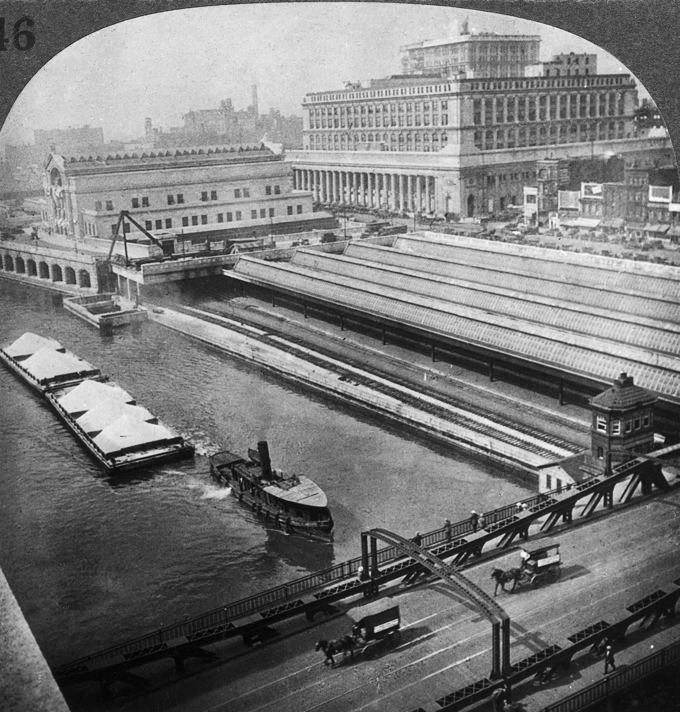 Black and white aerial photo of Chicago River canal with a tugboat pushing barges past a large train station and industrial buildings, with horse-drawn carriages on a nearby bridge.