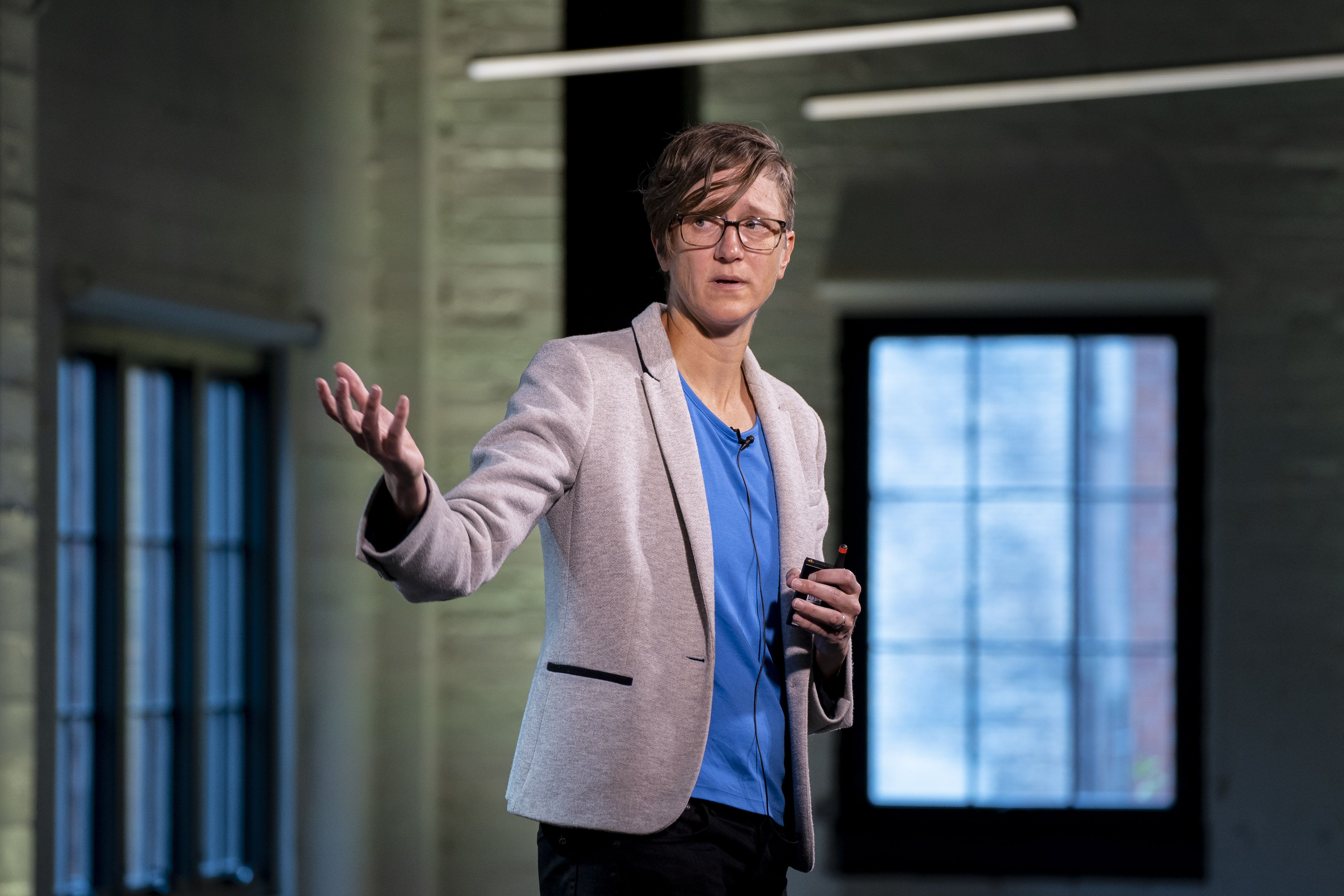 A woman in a light colored blazer speaks while holding out one of her hands.