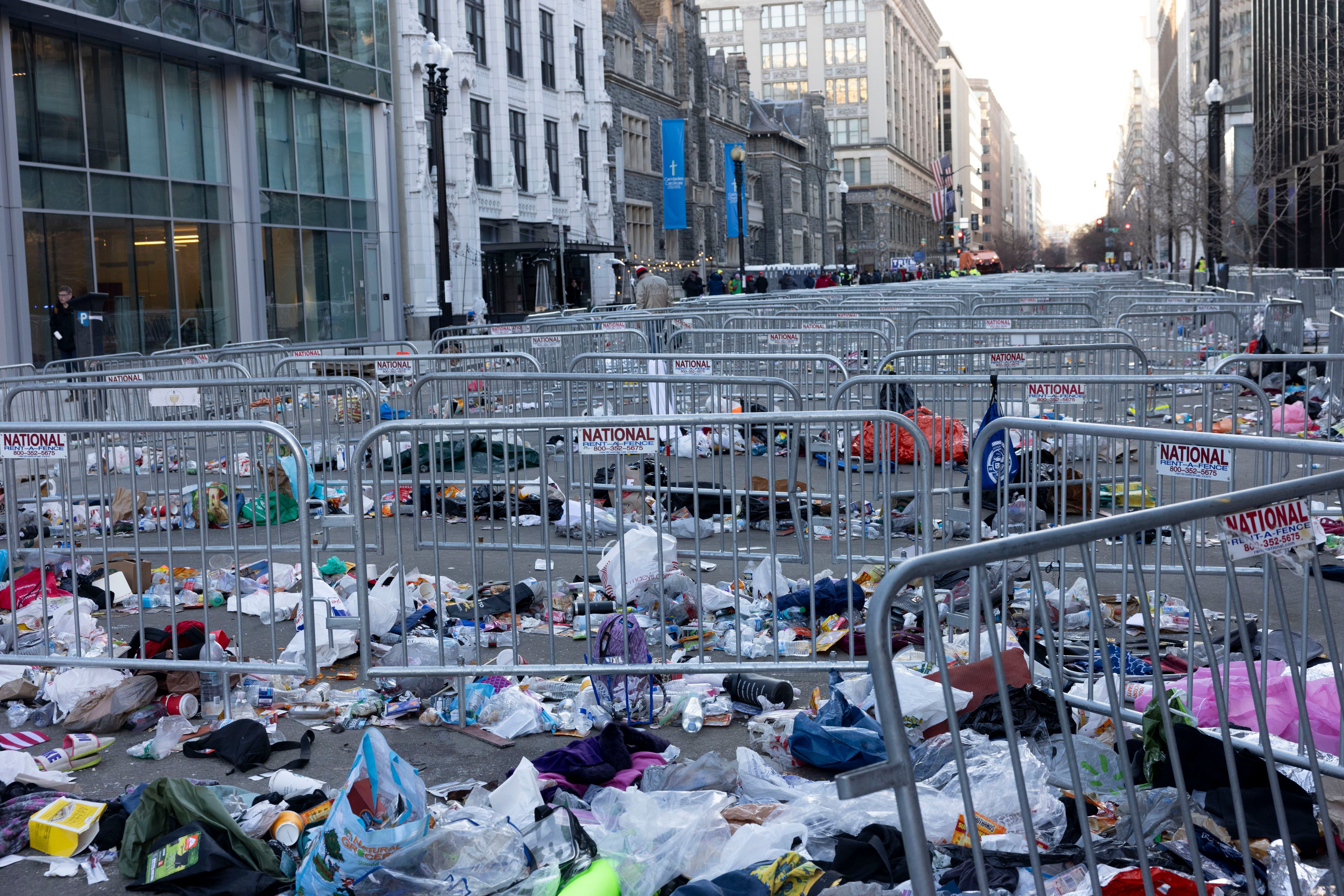 DC street fenced off with metal barricades labeled "National," littered heavily with trash including bags, bottles, and discarded items, between tall buildings under daylight.
