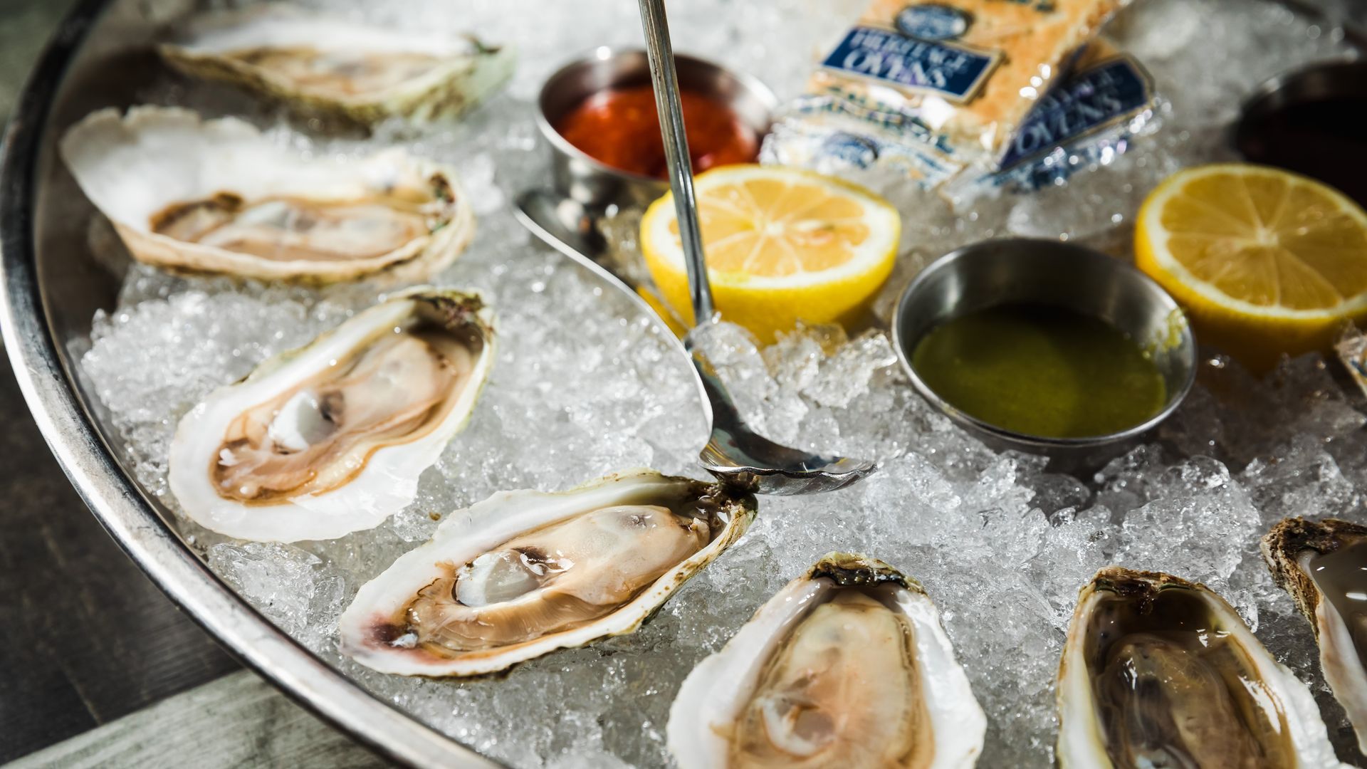 A metal tray on crushed ice holds fresh oysters on the half shell, bright lemon halves, and small metal bowls of sauces, creating a colorful, seaside seafood display.
