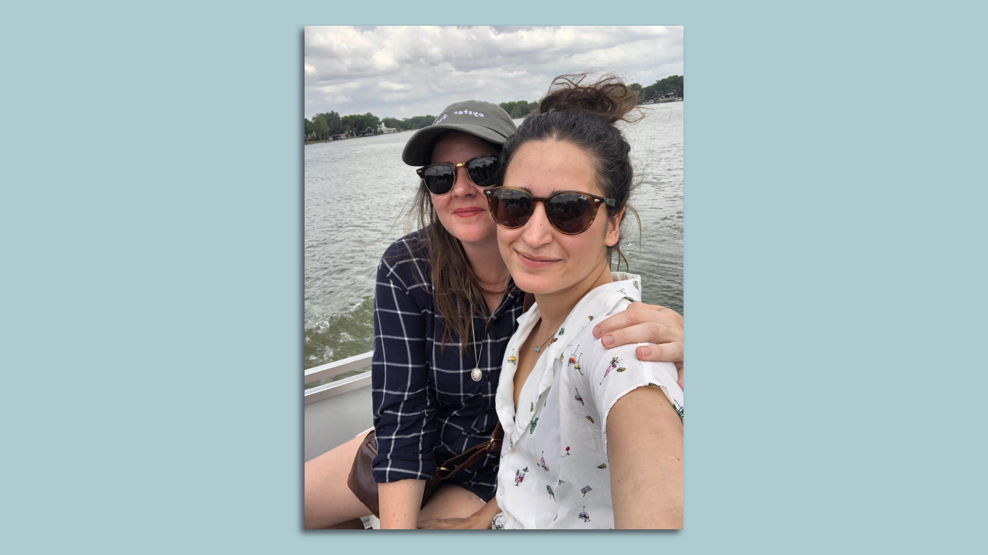Two smiling women, both wearing sunglasses, one in a dark shirt and hat and the other in a white blouse, sit in a boat with water behind them and trees in the distance. 